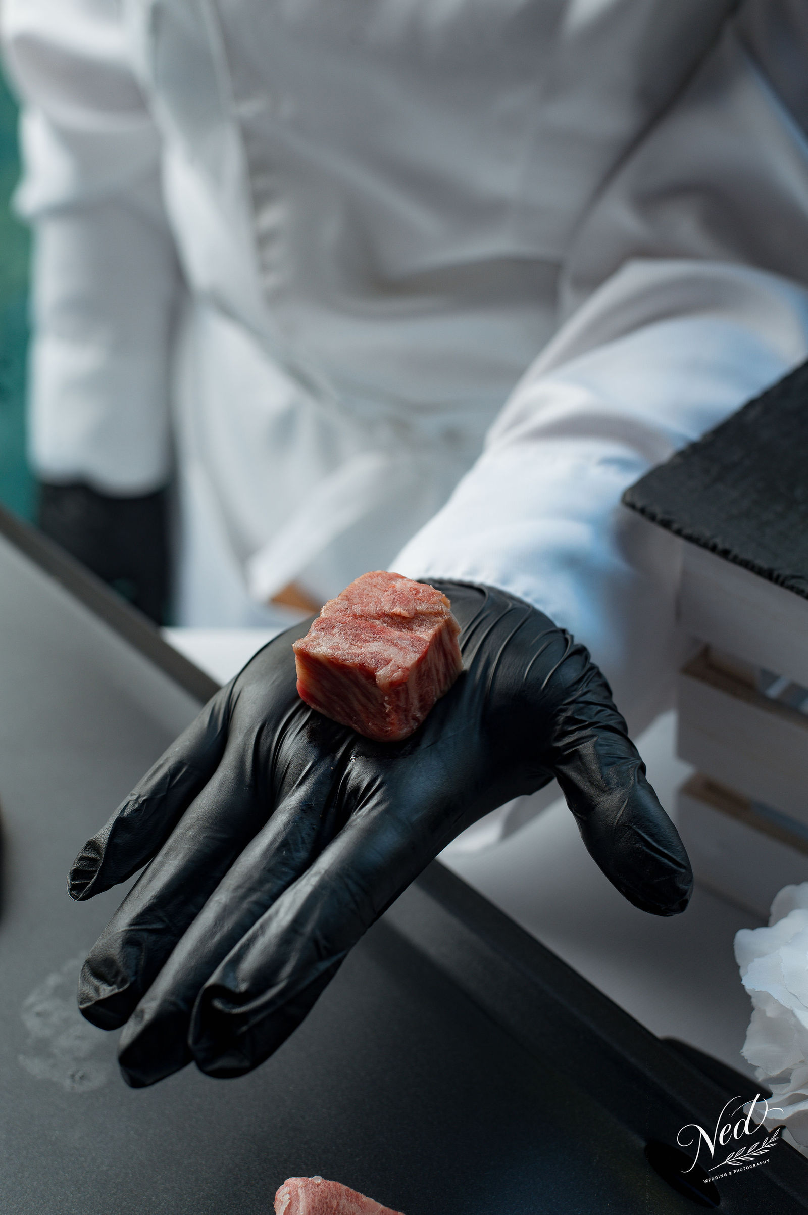 Main gantee de noir tenant un morceau de viande crue, personne en blouse blanche en arriere-plan