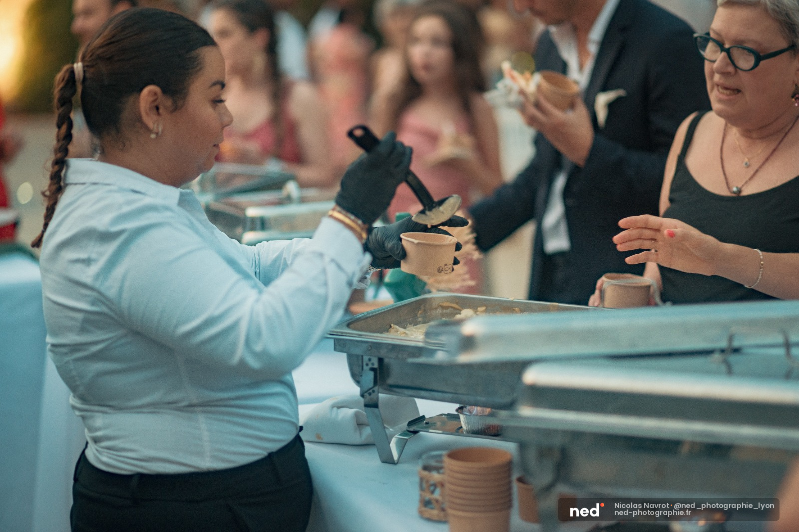 Femme en chemise blanche et gants noirs verse une preparation dans un gobelet devant des participants attentifs lors d'un atelier culinaire collectif