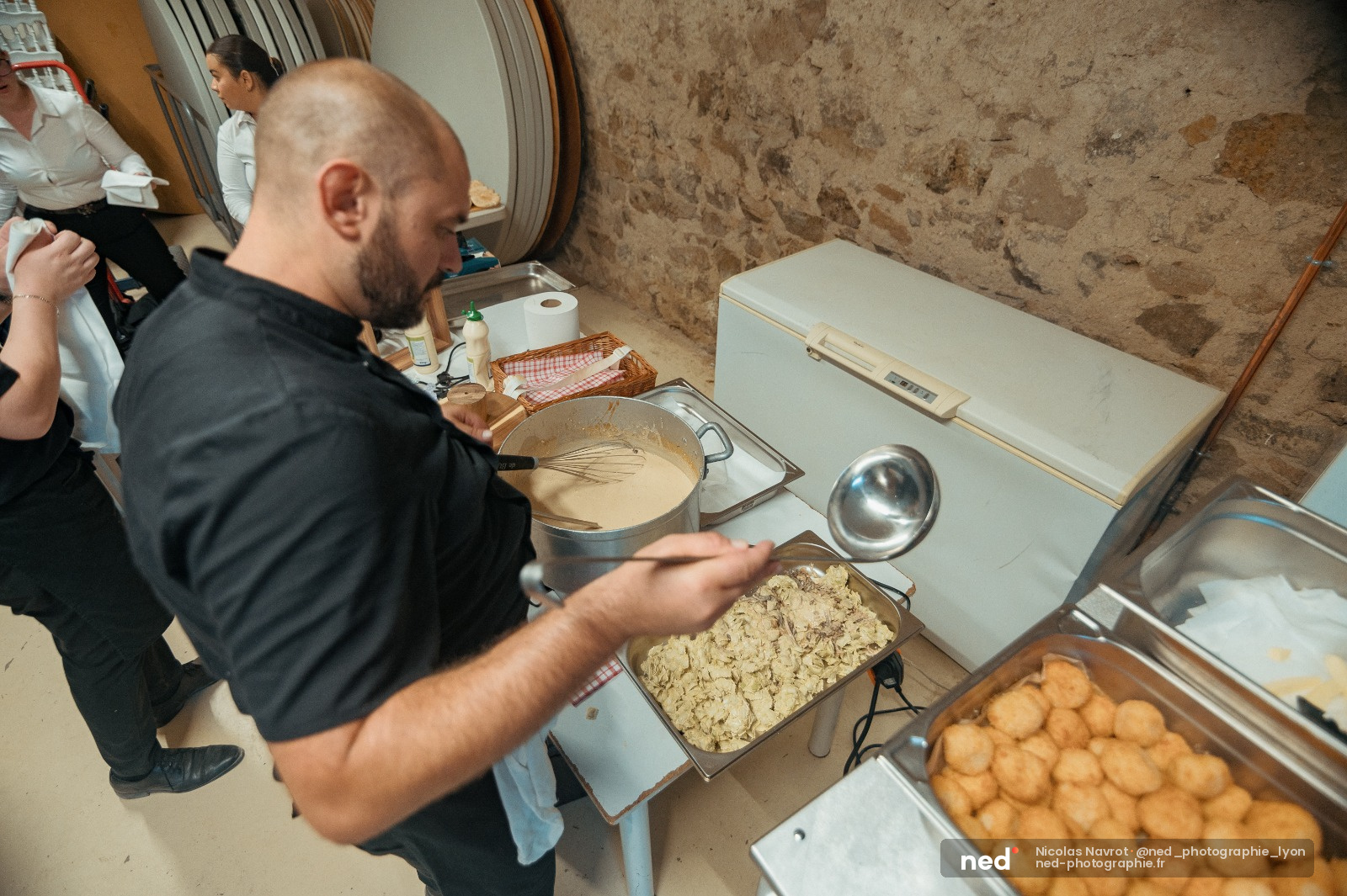 Homme en tee-shirt noir mélange une préparation culinaire dans un saladier, entouré de plats et participants en cuisine.
