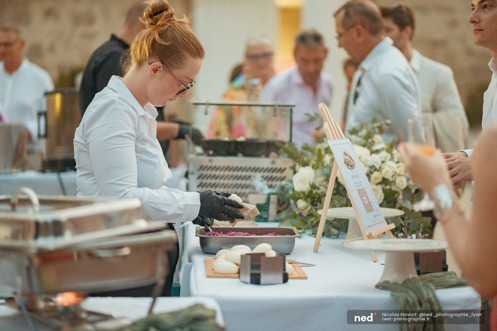 Femme en tablier blanc et gants noirs prépare un plat lors d'un atelier culinaire, participants observent en arrière-plan