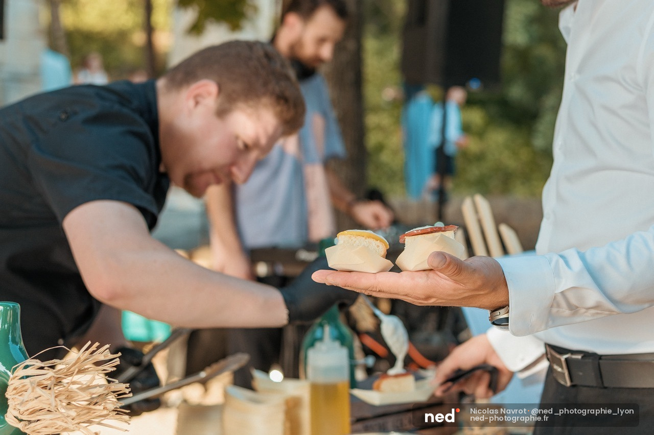 Formateur en chemise blanche presente des tartines a des participants assis autour d'une table en exterieur, sous les arbres