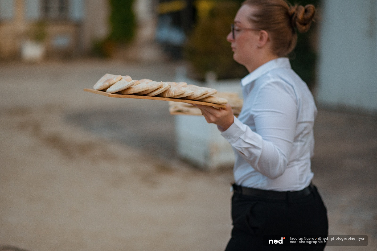 Femme en chemise blanche tenant une planche de baguettes fraîches devant une façade en pierre