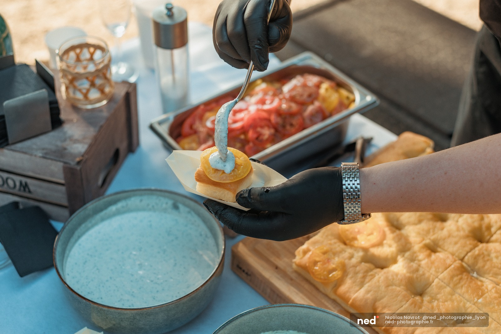 Chef en gants noirs dresse un canapé avec sauce bleue sur assiette blanche, table de travail avec ingredients et plats