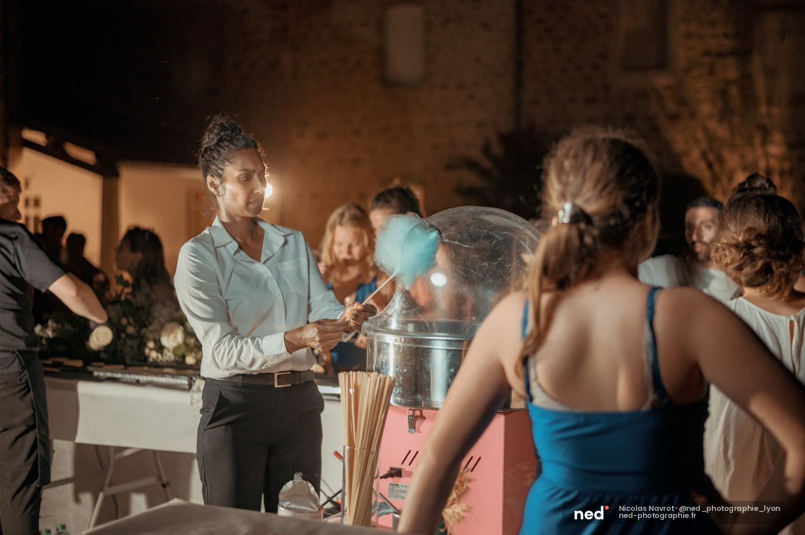 Serveuse en chemise blanche versant un cocktail bleu devant des convives en tenue de soirée, décor nocturne en pierre