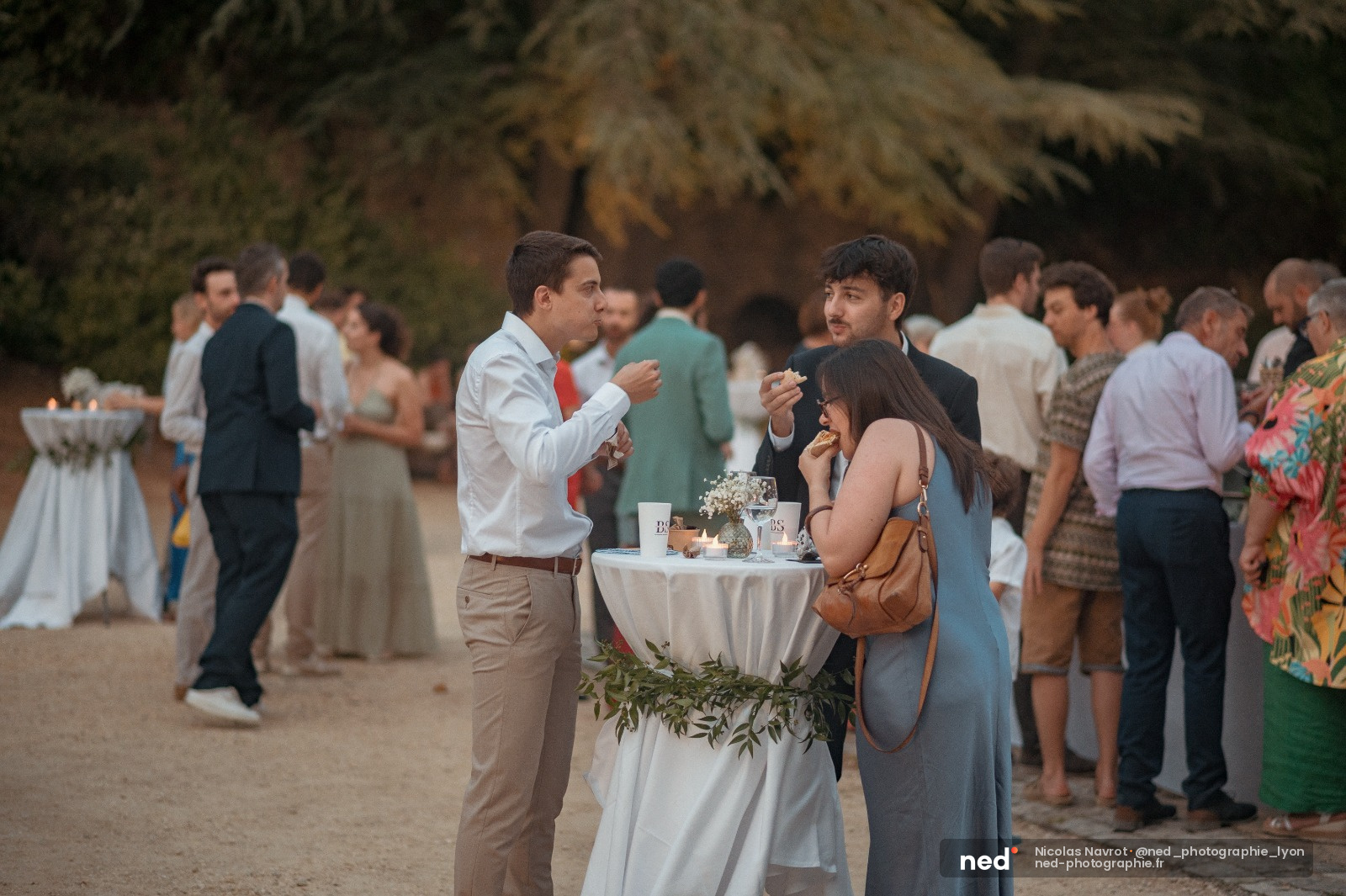 Invités discutant autour d'une table haute blanche lors d'un cocktail de mariage en extérieur, sous une arcature de végétation