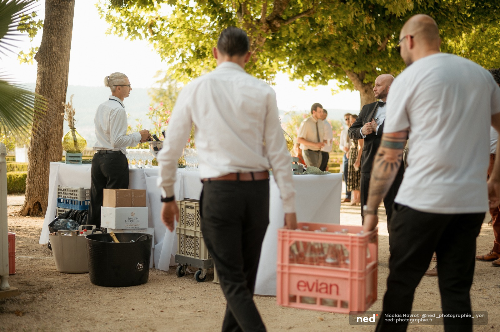 Hommes en costume blanc et noir discutent autour de tables de service en extérieur, sous des arbres avec vue sur l'eau.