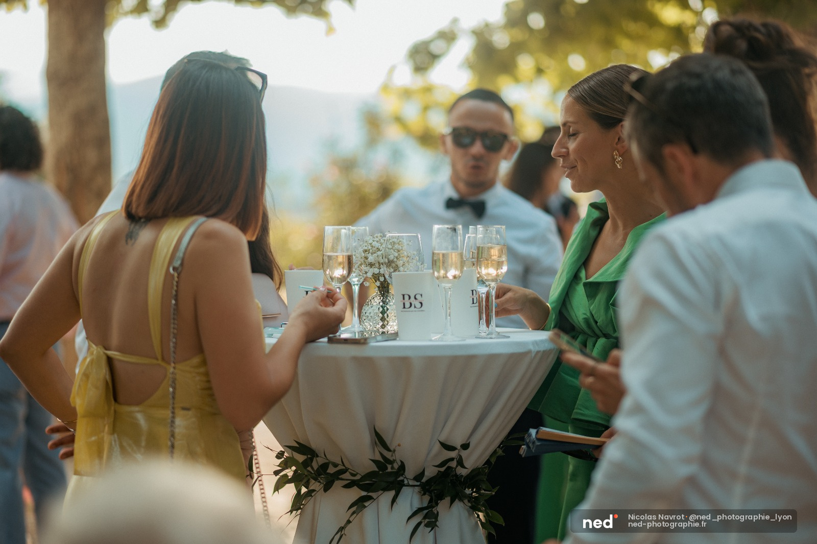 Invités en tenue élégante discutent autour d'une table blanche avec flûtes de champagne et fleurs blanches, sous les arbres en extérieur.