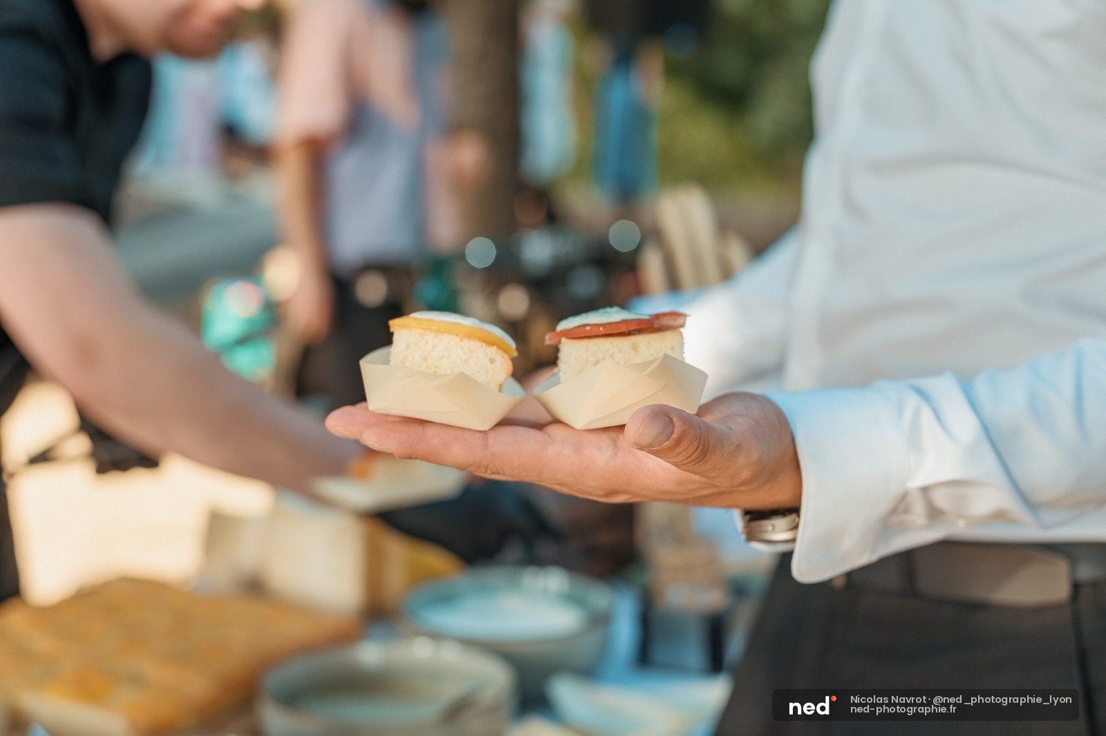 Main tenant deux portions de fromage blanc avec croute doree, groupe de personnes autour en exterieur par beau temps