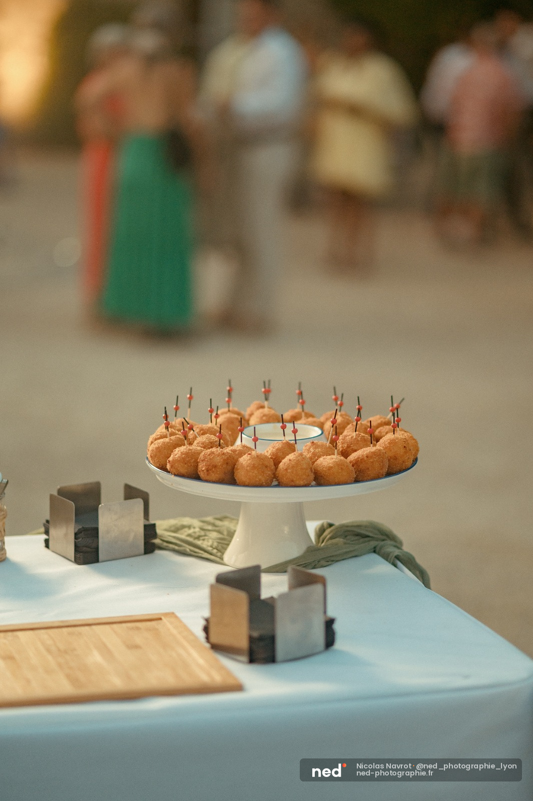 Présentoir blanc avec boulettes dorées piquées de petits drapeaux, sur table blanche décorée, invités flous en arrière-plan
