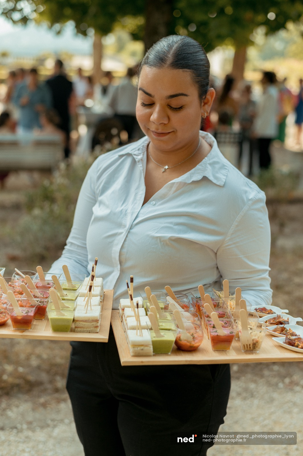 Femme en chemise blanche présente des verrines et canapés sur plateau en bois lors d'un événement extérieur