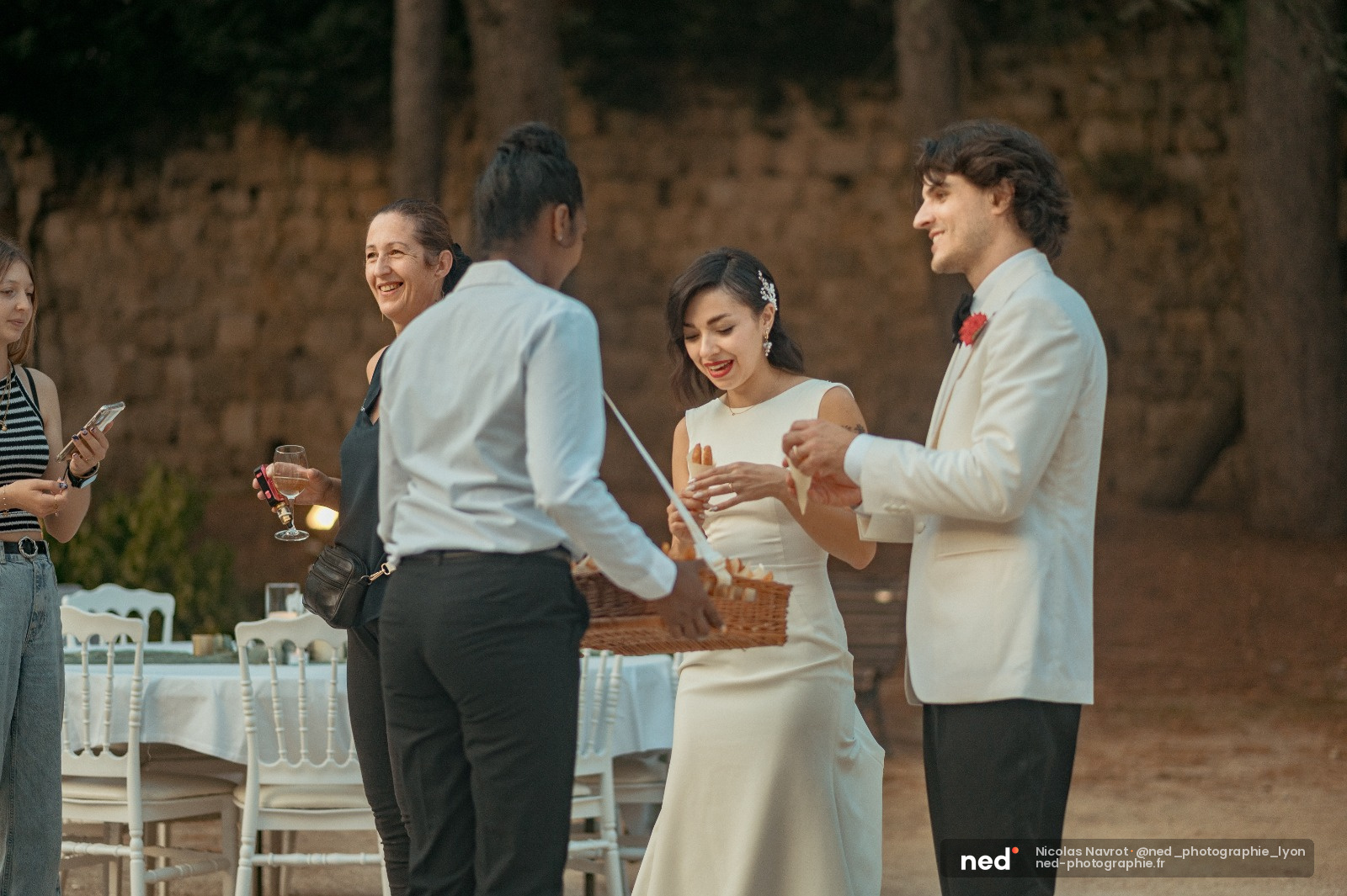 Mariee en robe blanche et marie en costume clair discutent avec des invites pendant la reception en exterieur pres d'une vieille batisse