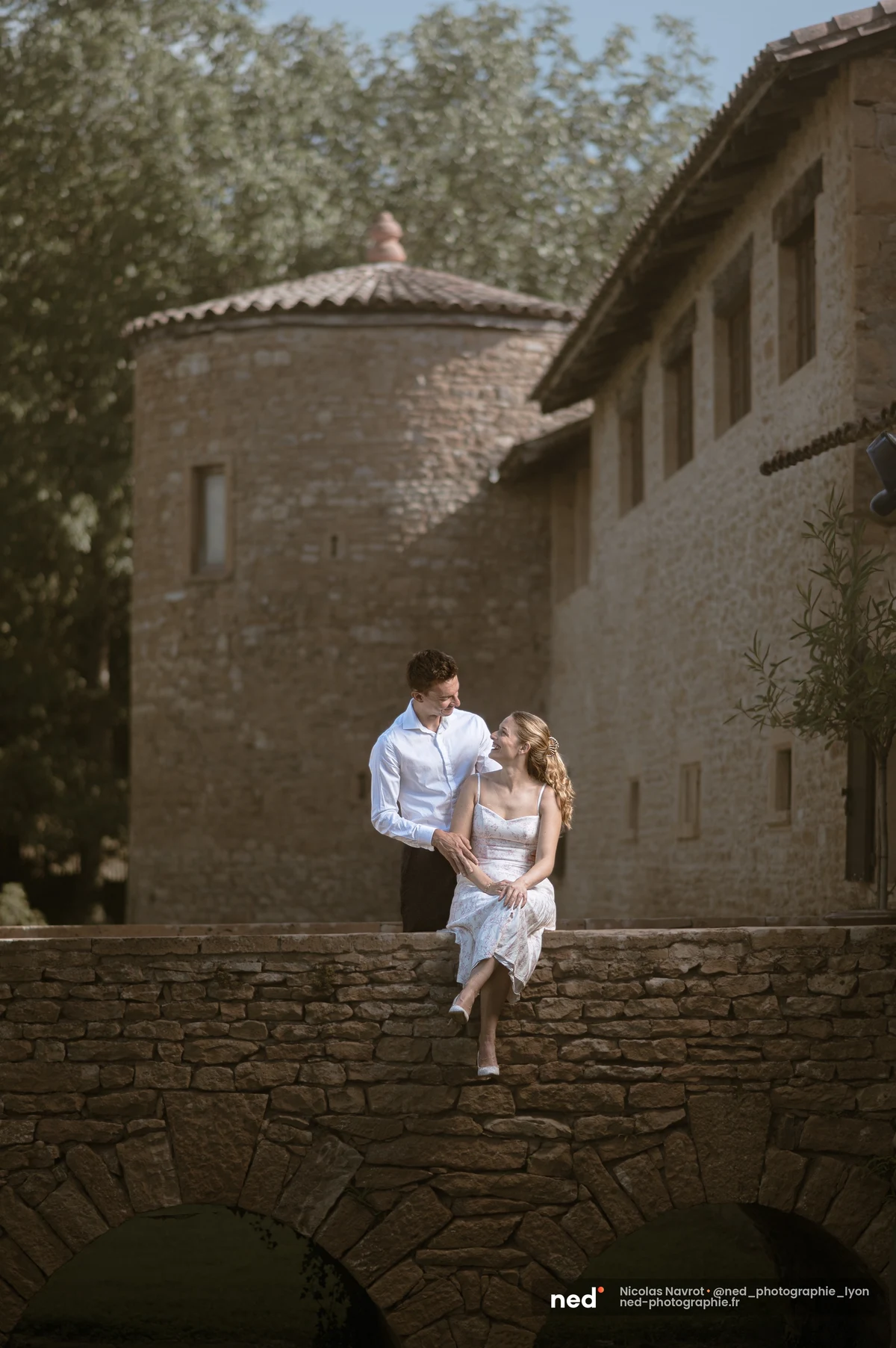 Femme assise sur un muret de pont en pierre, homme debout, tour de domaine derrière., séance photo de couple au Château du Sou