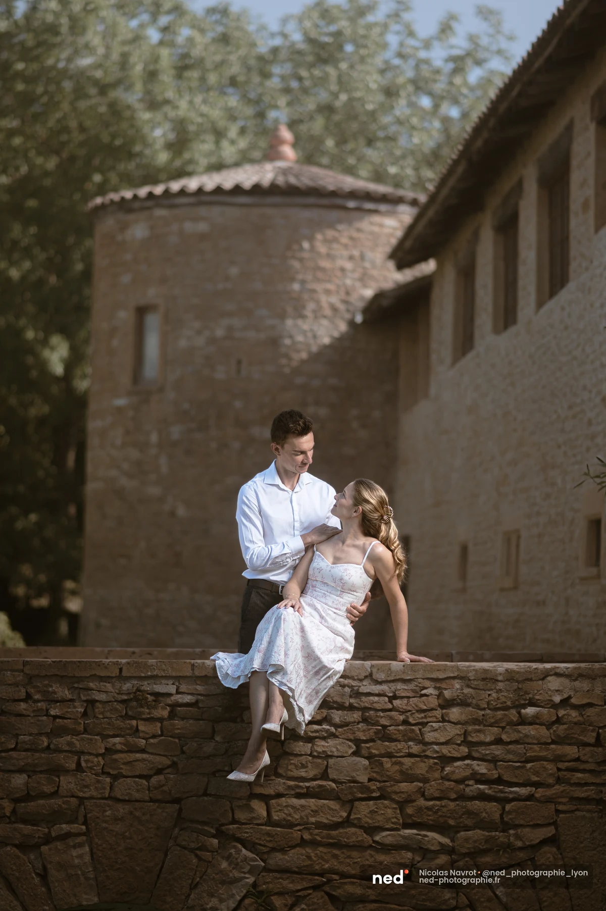 Couple assis sur un muret de pierre devant une tour ancienne, séance photo de couple au Château du Sou