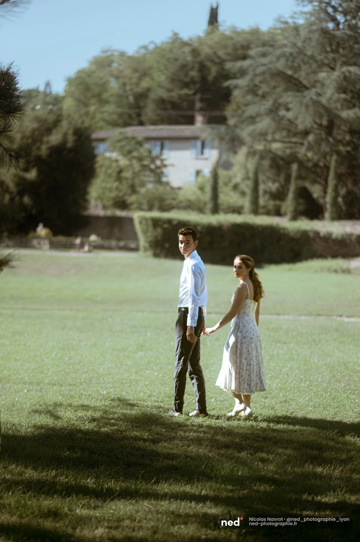Couple se tenant la main marchant sur une pelouse verte de domaine, séance photo de couple au Château du Sou