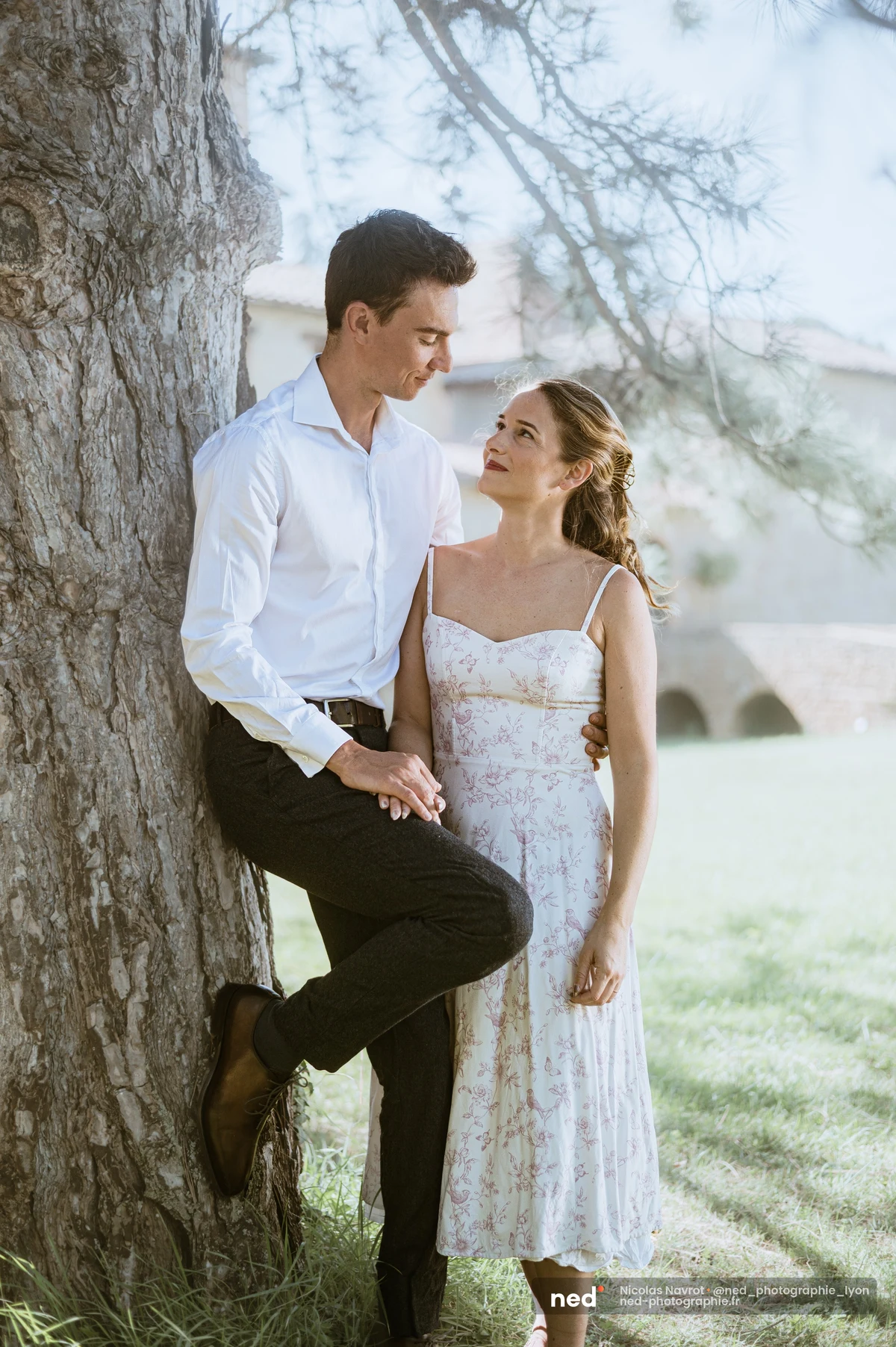Couple enlacé contre un pin, domaine en pierre en arrière-plan, lumière estivale., séance photo de couple au Château du Sou