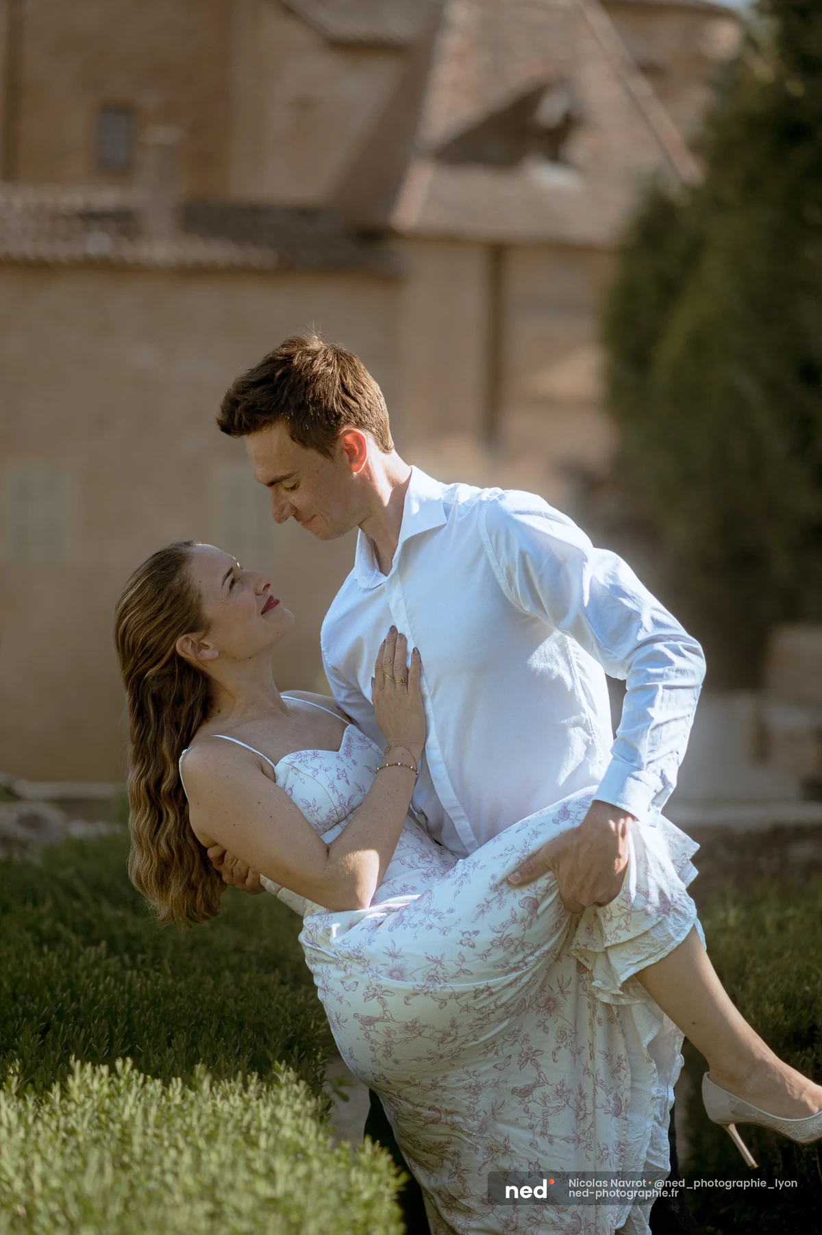 Homme soulevant sa compagne en robe fleurie devant un château en pierre, séance photo de couple au Château du Sou