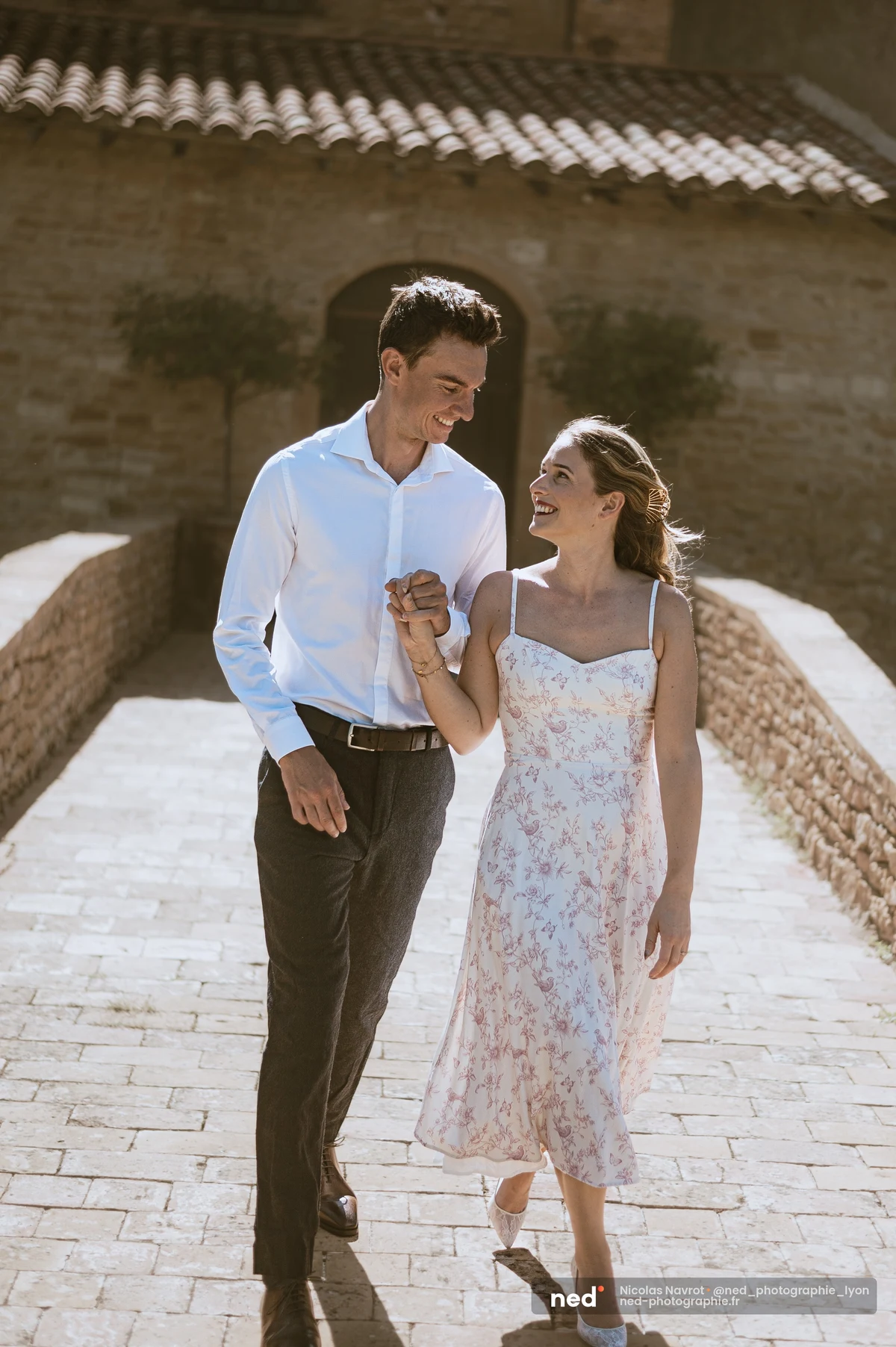 Couple marchant main dans la main sur une allée pavée, mas en pierre dorée, soleil., séance photo de couple au Château du Sou