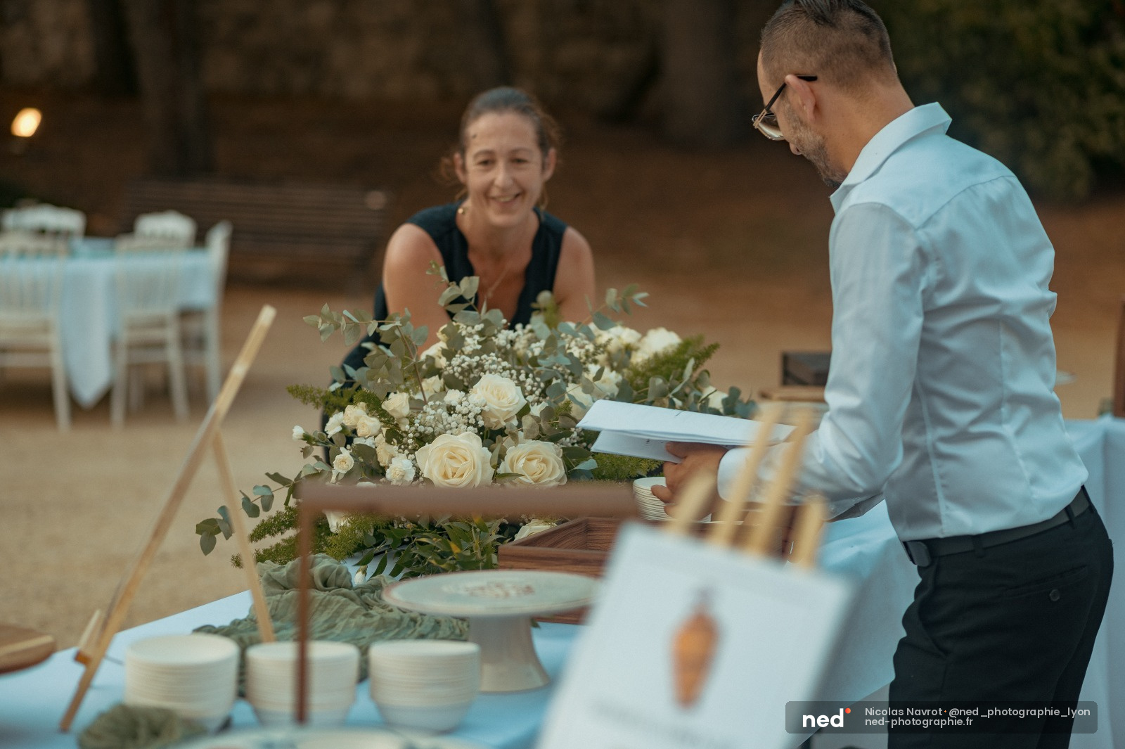 Homme en chemise bleue consulte ses notes près d'une table décorée de fleurs blanches et de verdure, femme sourit en arrière-plan lors d'une cérémonie de mariage.