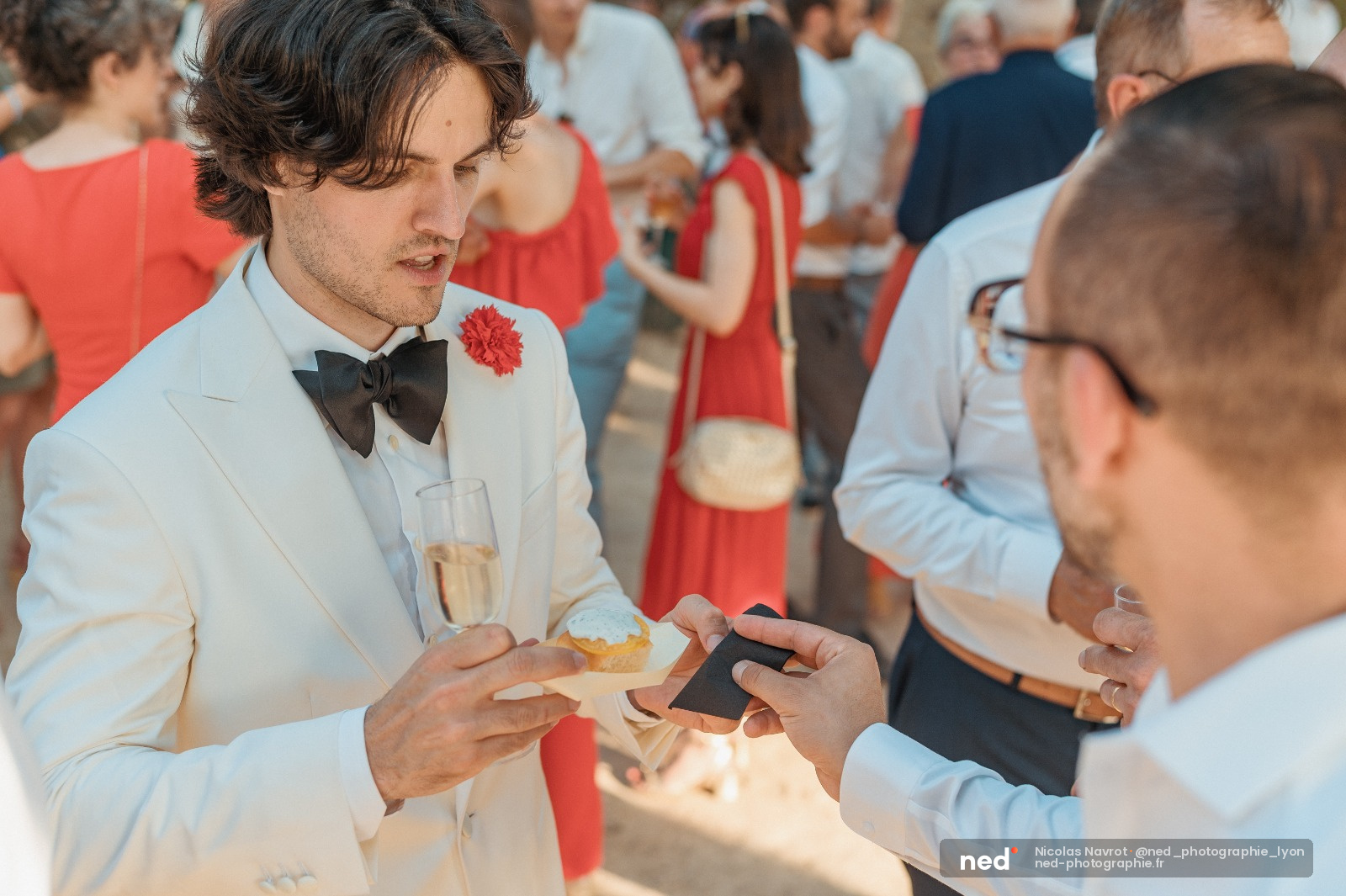 Homme en costume crème et nœud papillon noir discute avec un invité lors d'une réception de mariage en plein air