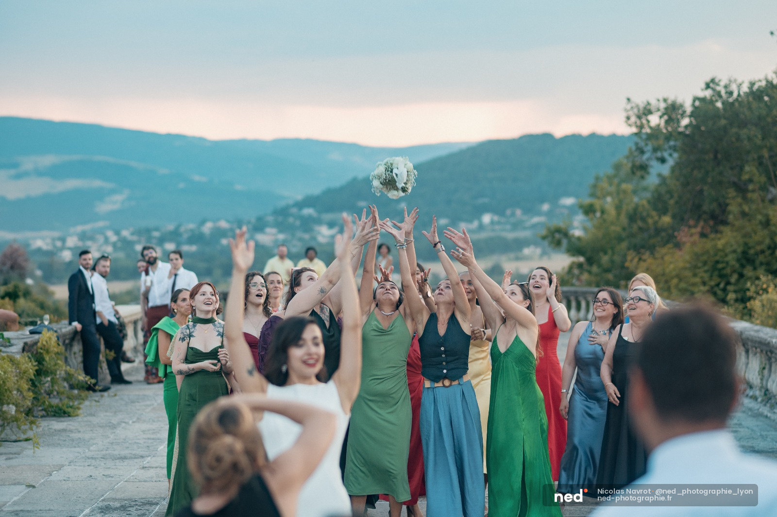 Groupe de femmes en robes colorées lèvent les bras pour attraper le bouquet de mariée, montagnes verdoyantes en arrière-plan