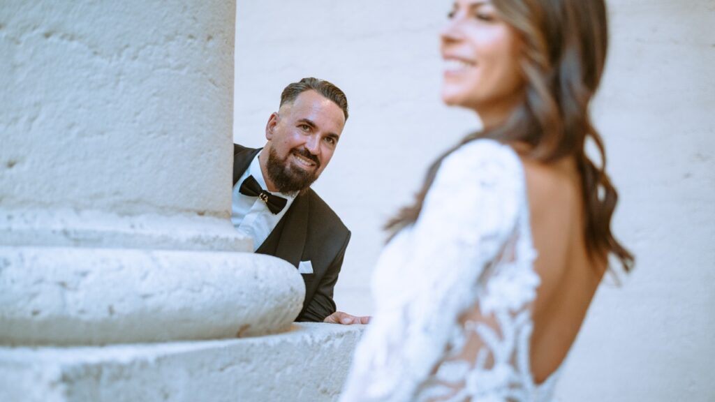 Marié en costume et nœud papillon regardant sa fiancée en robe blanche derrière une colonne blanche