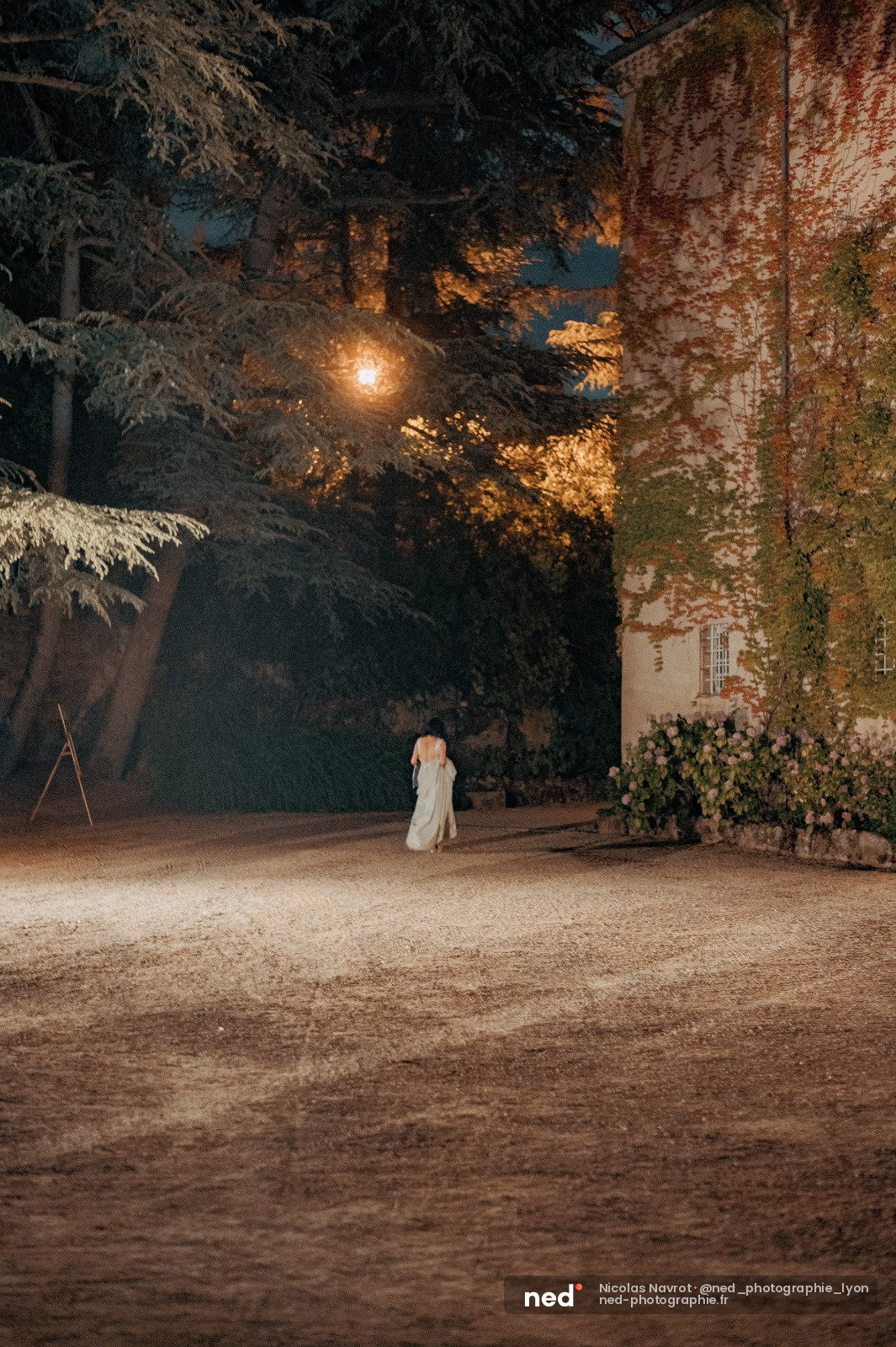 Mariée en robe blanche debout seule dans une cour pavée, eclairée par un lampadaire la nuit, murs en pierre couverts de lierre