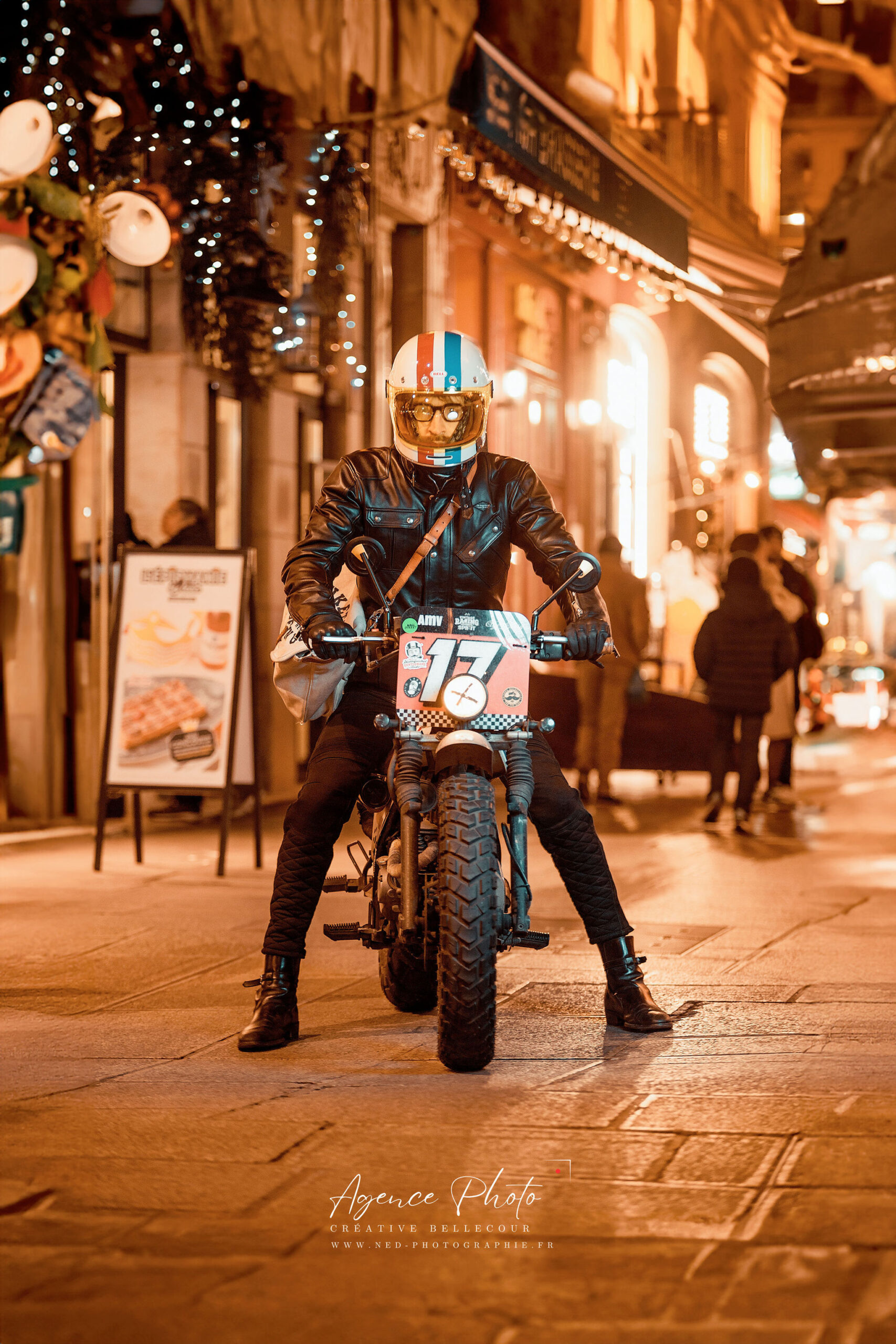 Motard au casque rétro et blouson noir sur petite moto de course, rue commerçante illuminée avec guirlandes et boules lumineuses