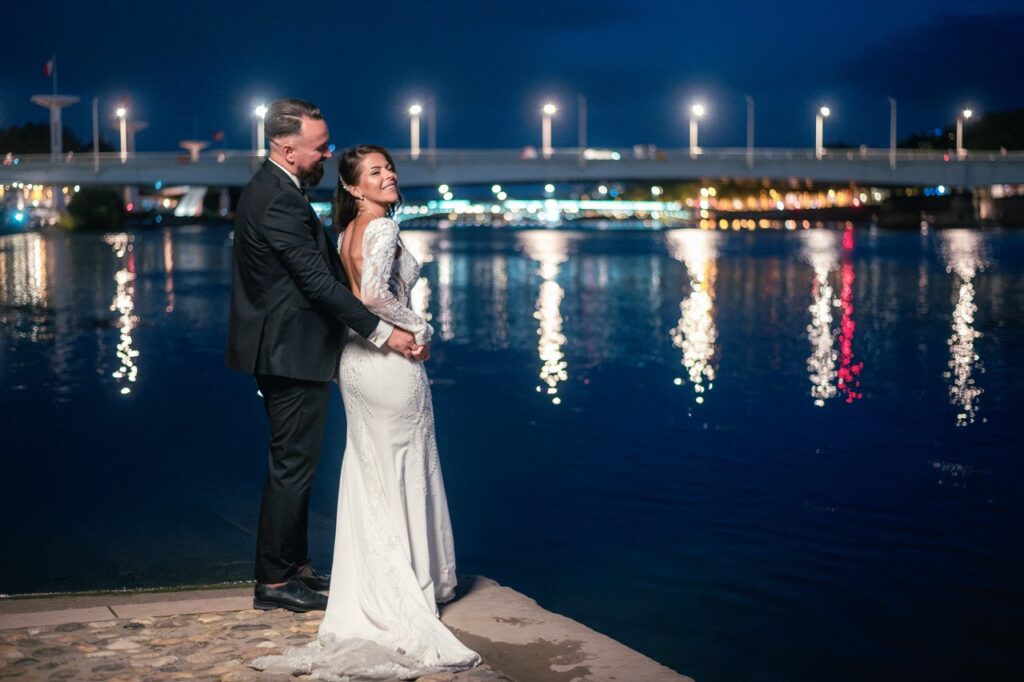 Mariée en robe blanche et marié en costume noir au bord du Rhône la nuit, pont illuminé en arrière-plan