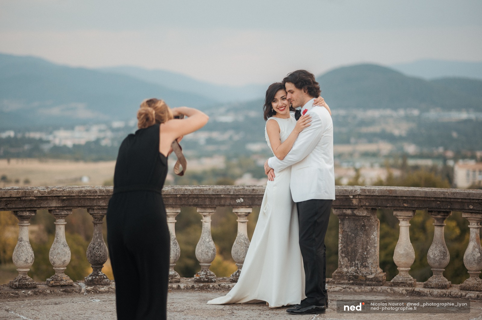 Couple enlacé en robe et costume blanc sur une terrasse en pierre, femme photographe en noir au premier plan, vallée et montagnes en arrière-plan
