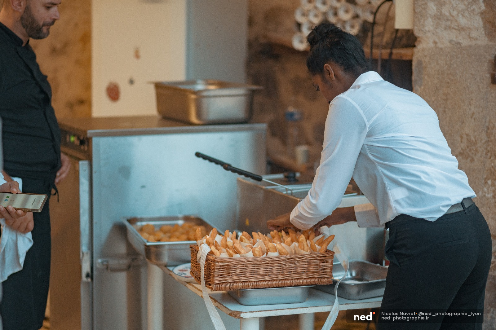 Photographe en chemise blanche prenant des photos d'un buffet avec panier de viennoiseries en osier, homme en noir observant à gauche
