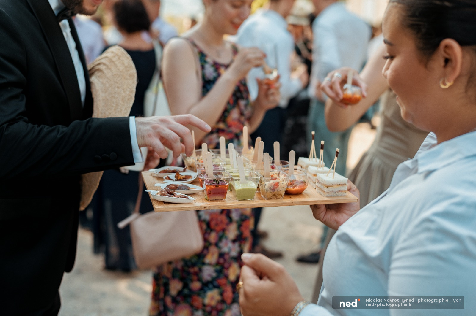 Invités en tenue habillée dégustant des amuse-bouches lors d'un cocktail de mariage en extérieur, plateau de canapés en premier plan