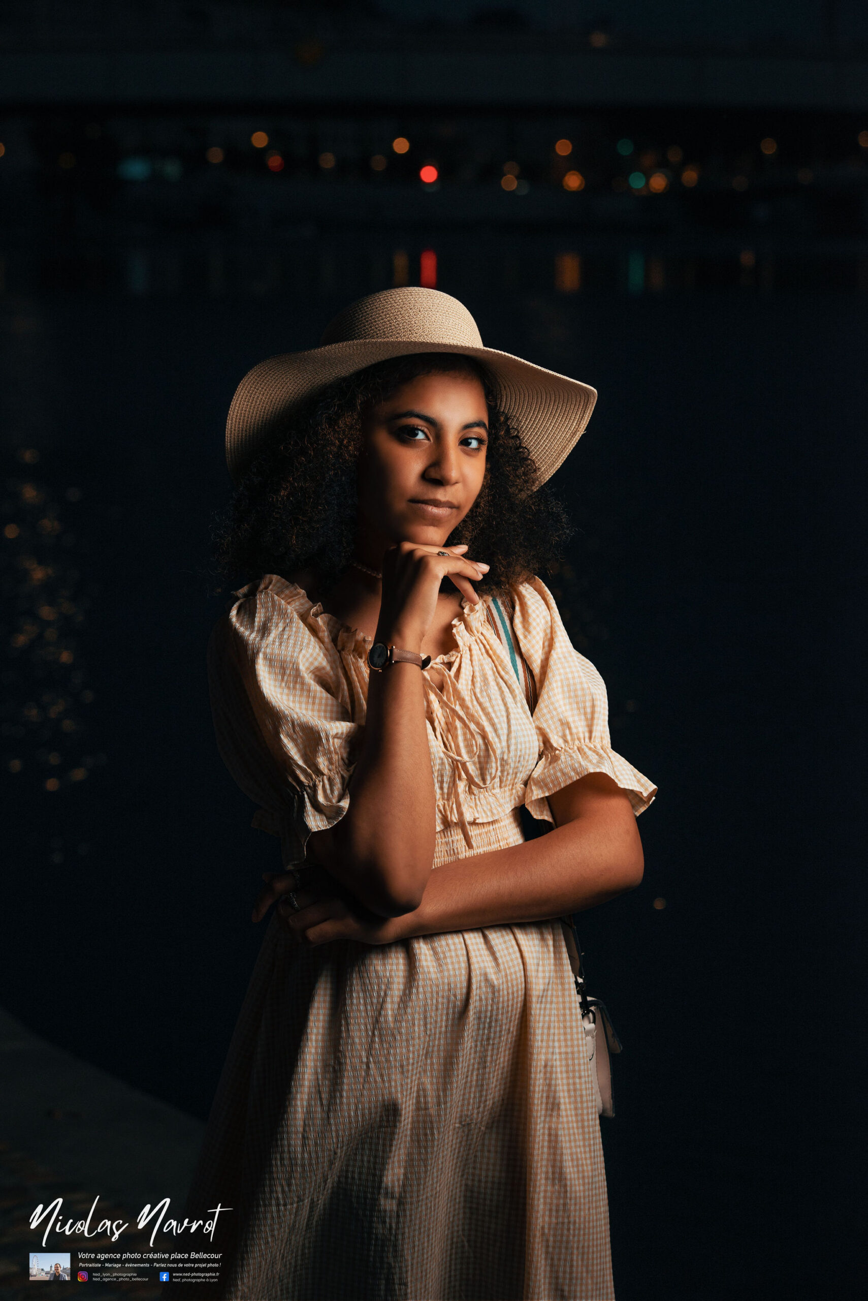 Jeune femme en robe pêche et chapeau paille regardant l'objectif, main au menton, fond noir avec bokeh de lumières urbaines