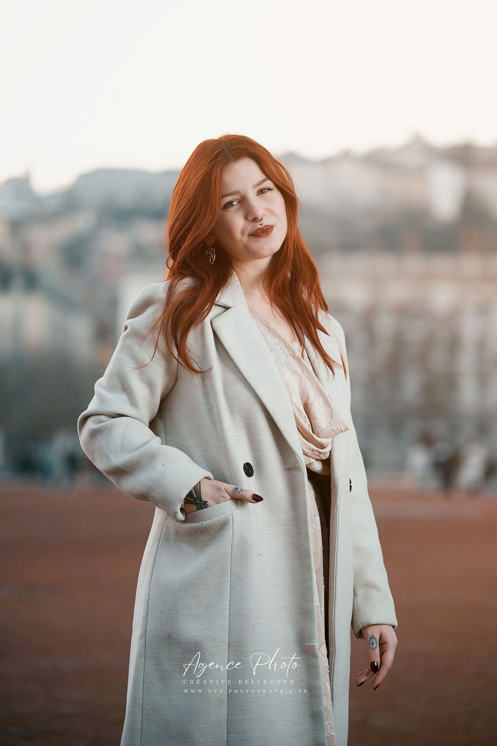 Femme aux cheveux roux portant un blazer beige clair, debout en extérieur face à un paysage urbain flou en arrière-plan