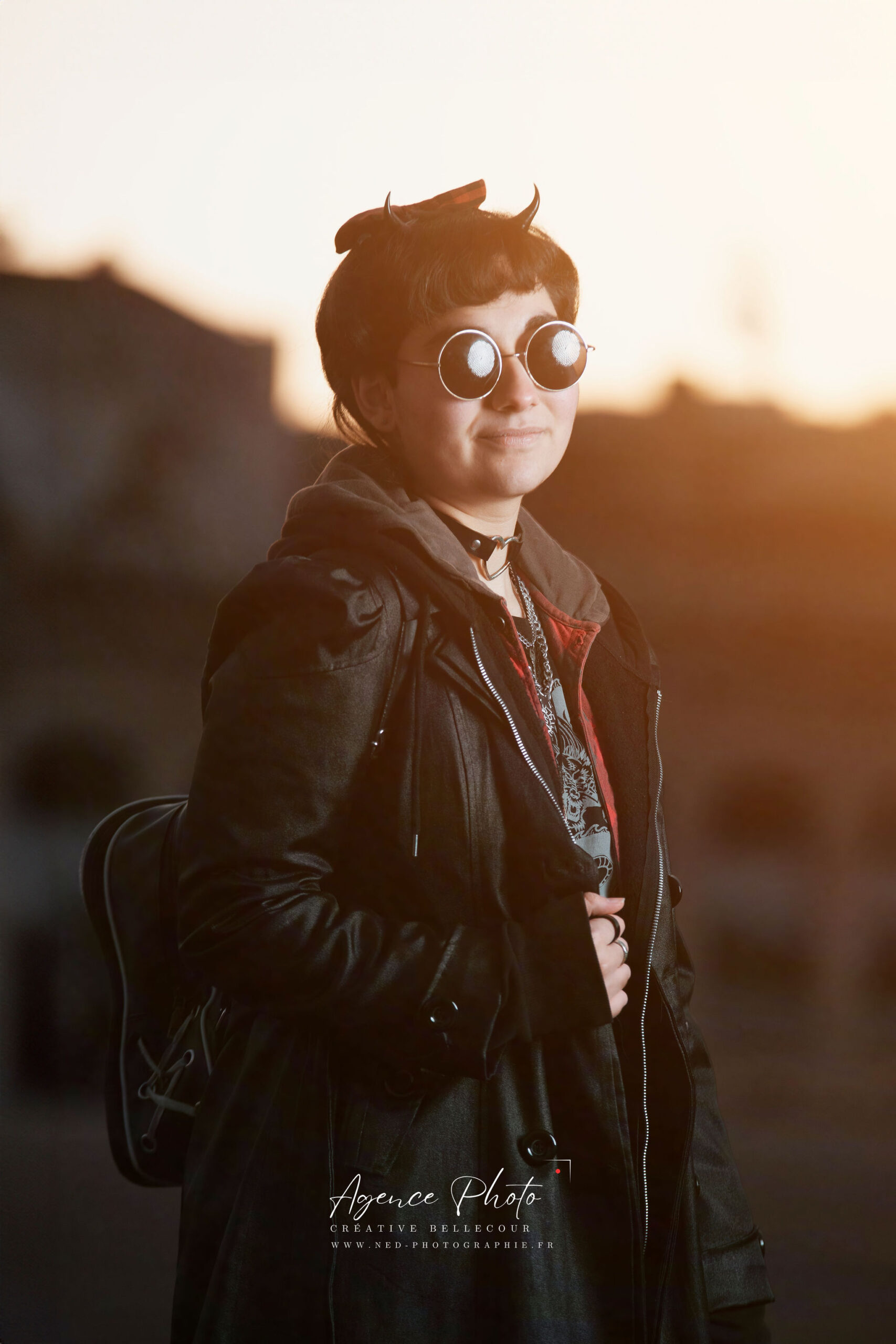 Femme aux cheveux courts et lunettes rondes porte un blouson noir, regardant vers l'arrière en extérieur au coucher de soleil