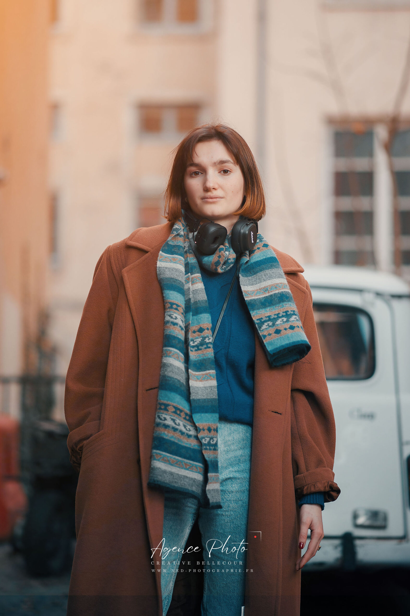 Jeune femme en manteau terracotta et écharpe bleue posant devant des immeubles en pierre, casque audio autour du cou
