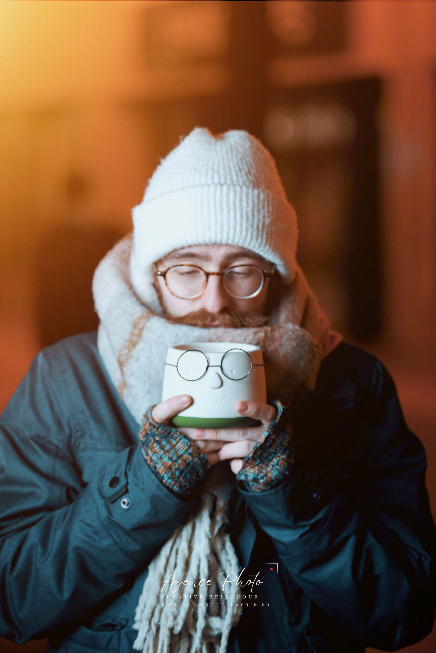 Homme avec lunettes, bonnet blanc et écharpe tient une tasse blanche devant son visage sur fond architectural orange.