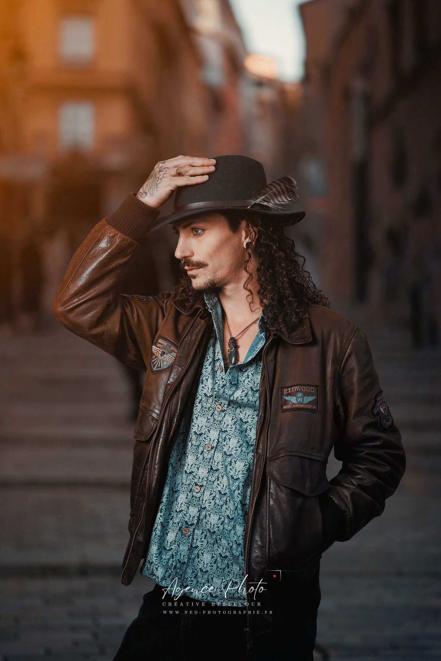 Homme aux cheveux longs ajuste son chapeau noir, veste cuir marron, chemise bleu-vert, en ruelle pavée avec facades en arrière-plan