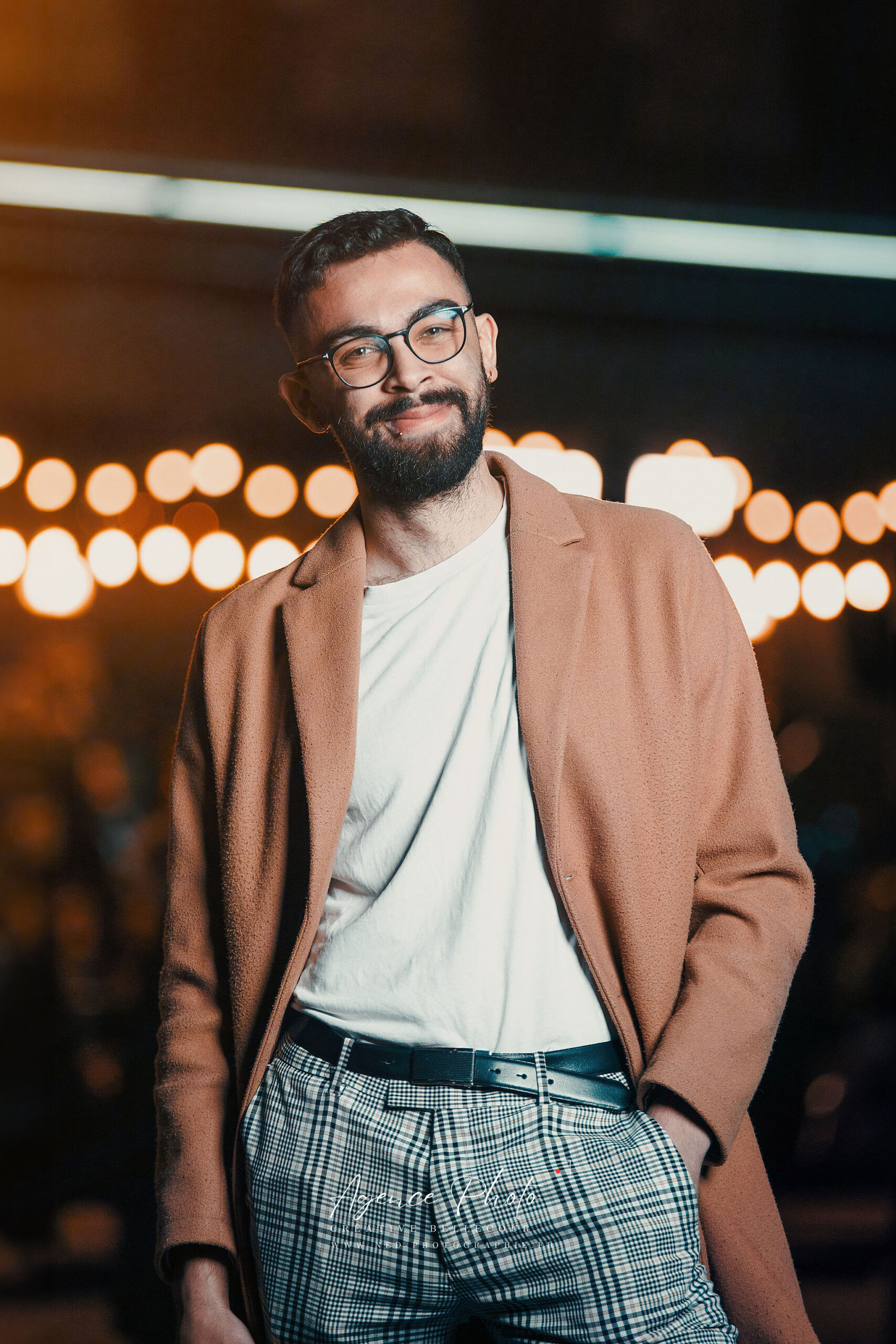 Homme barbu portant des lunettes, veste camel et pantalon carreaux, posant devant des lumières de ville