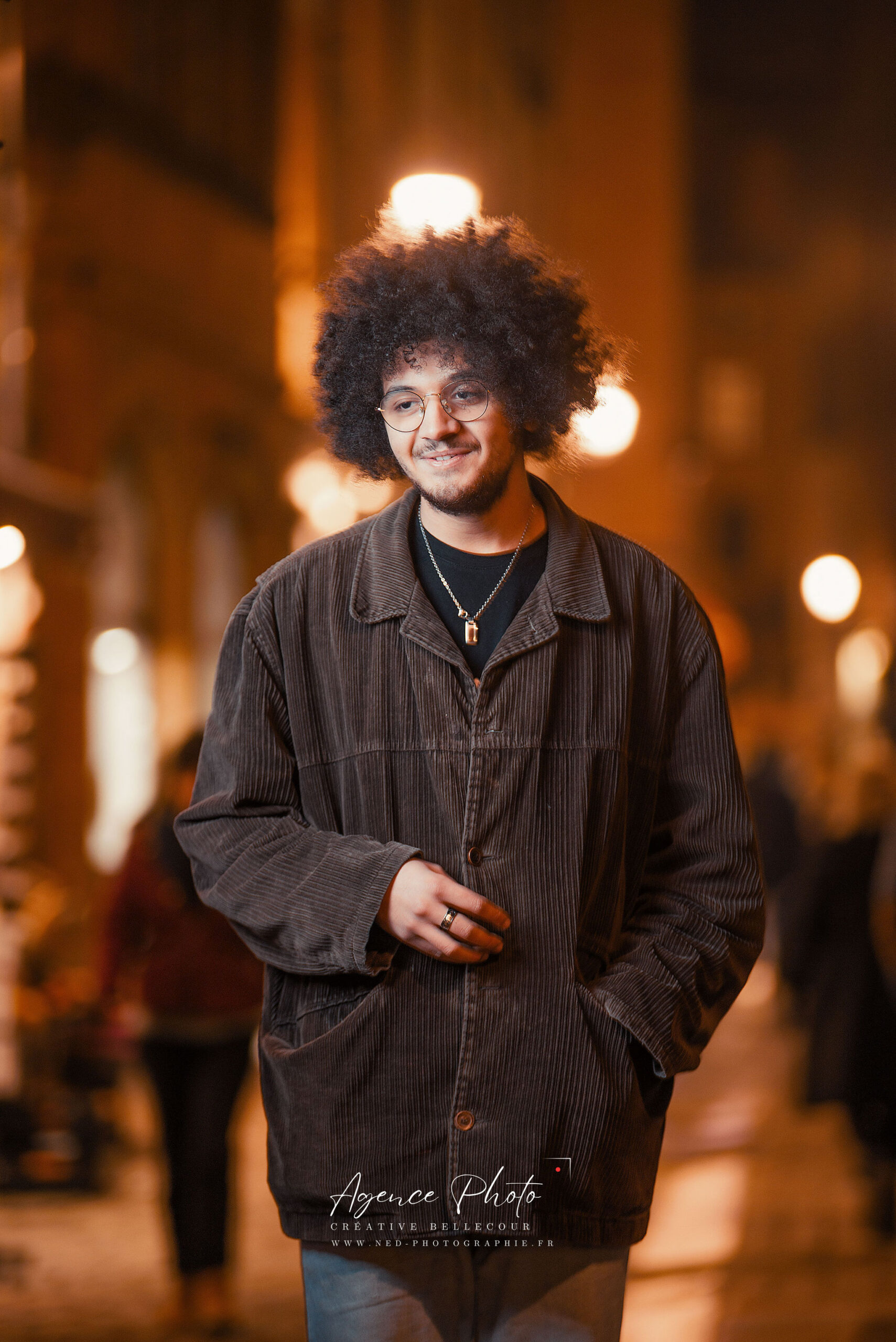 Jeune homme aux cheveux afro et lunettes, veste noire à rayures, sourit face à la caméra dans une rue illuminée en soirée