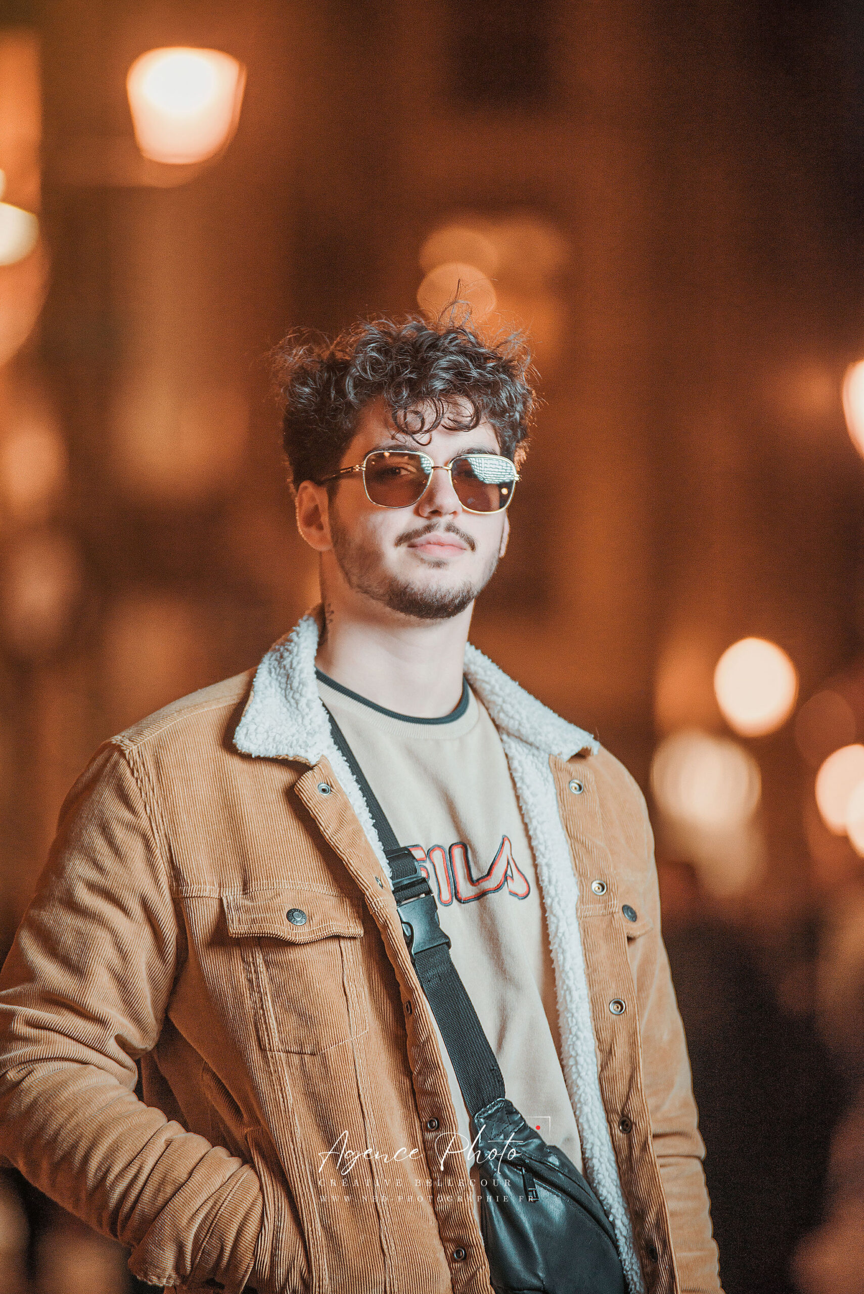 Jeune homme en veste marron et lunettes de soleil pose devant des lumières de nuit en ville