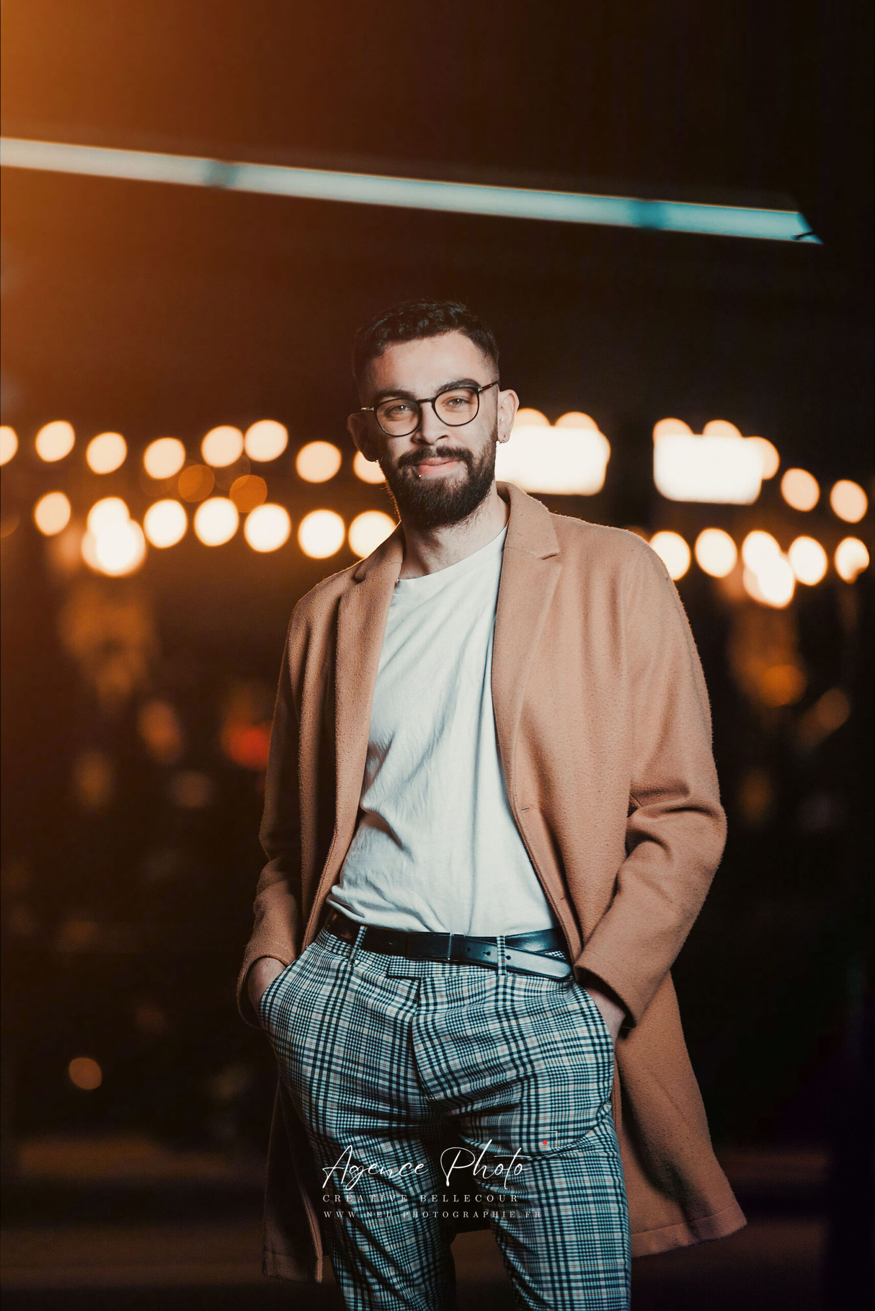 Homme avec lunettes en blazer beige et pantalon carreaux bleu, mains dans les poches, devant lumieres de ville floues