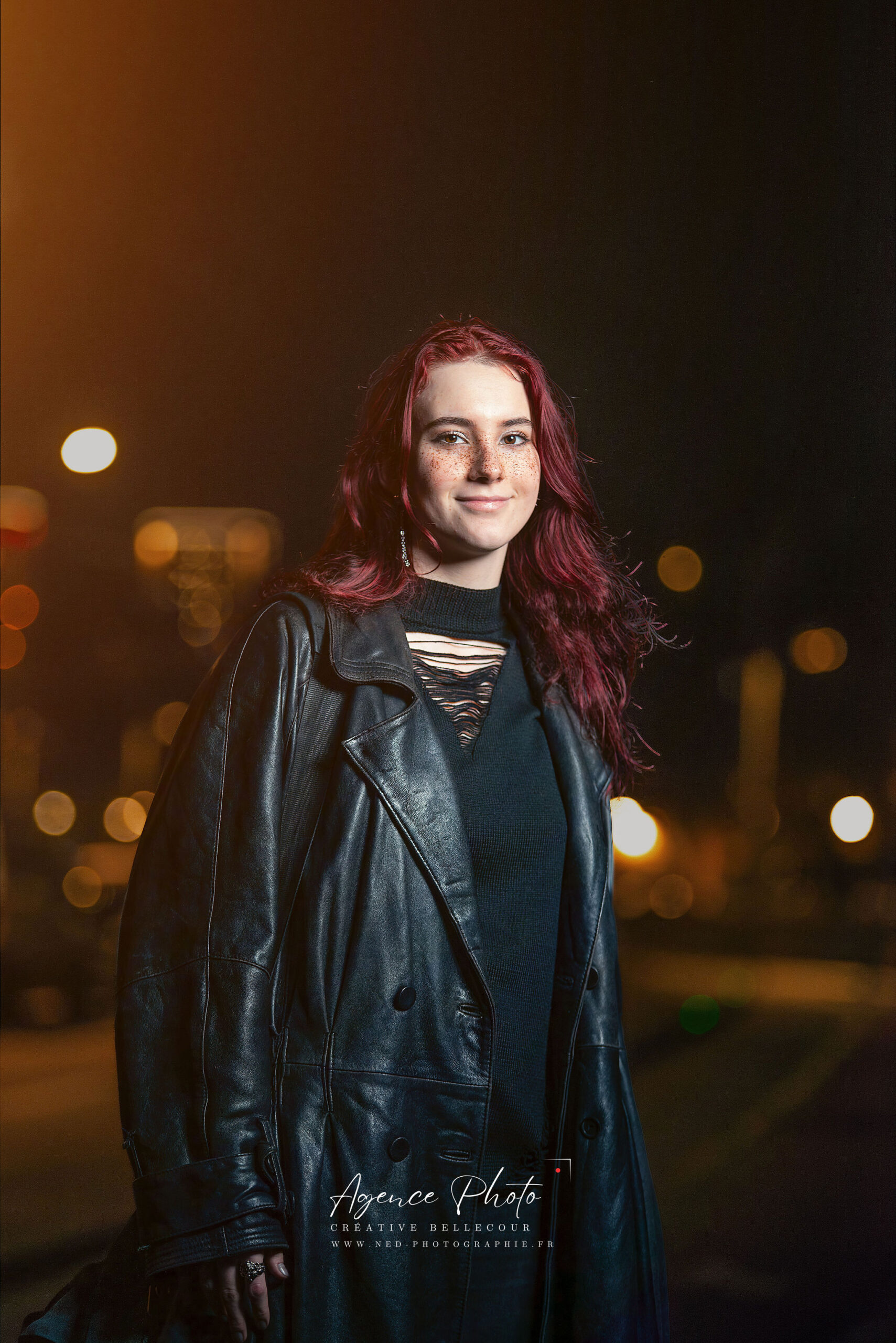 Jeune femme aux cheveux rouges sourit face à la caméra, veste noire en cuir, lumières de ville floues en arrière-plan