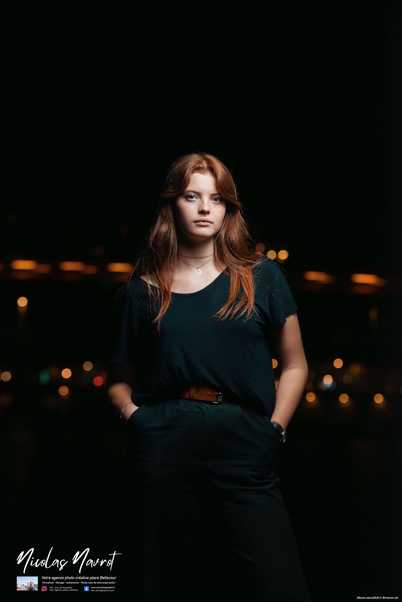 Jeune femme aux cheveux roux vêtue de noir, mains dans les poches, lumière des immeubles floutée en arrière-plan