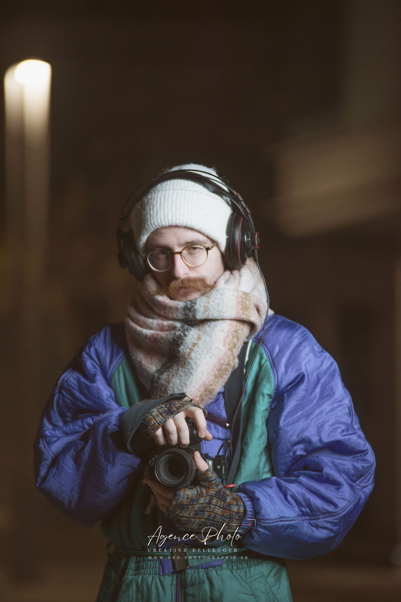 Homme barbu avec bonnet blanc et lunettes tient un appareil photo, casque sur les oreilles, veste bleue et verte en intérieur