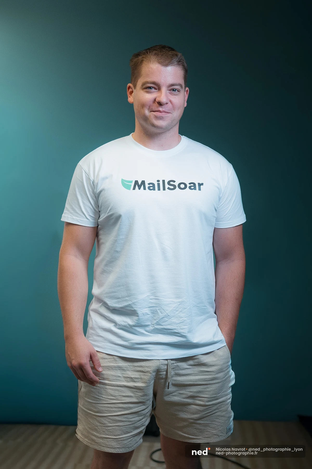 Homme debout en t-shirt blanc frappé du logo MailSoar et short beige, sur fond vert canard, mains derrière le dos.