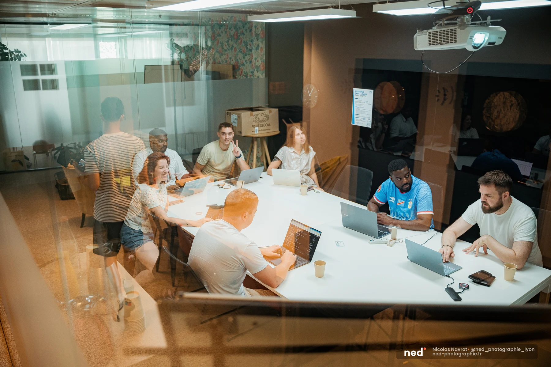 Vue à travers une vitre : groupe de personnes réunies autour d'une grande table blanche avec laptops, l'une d'elles debout se penche vers une collègue qui rit.