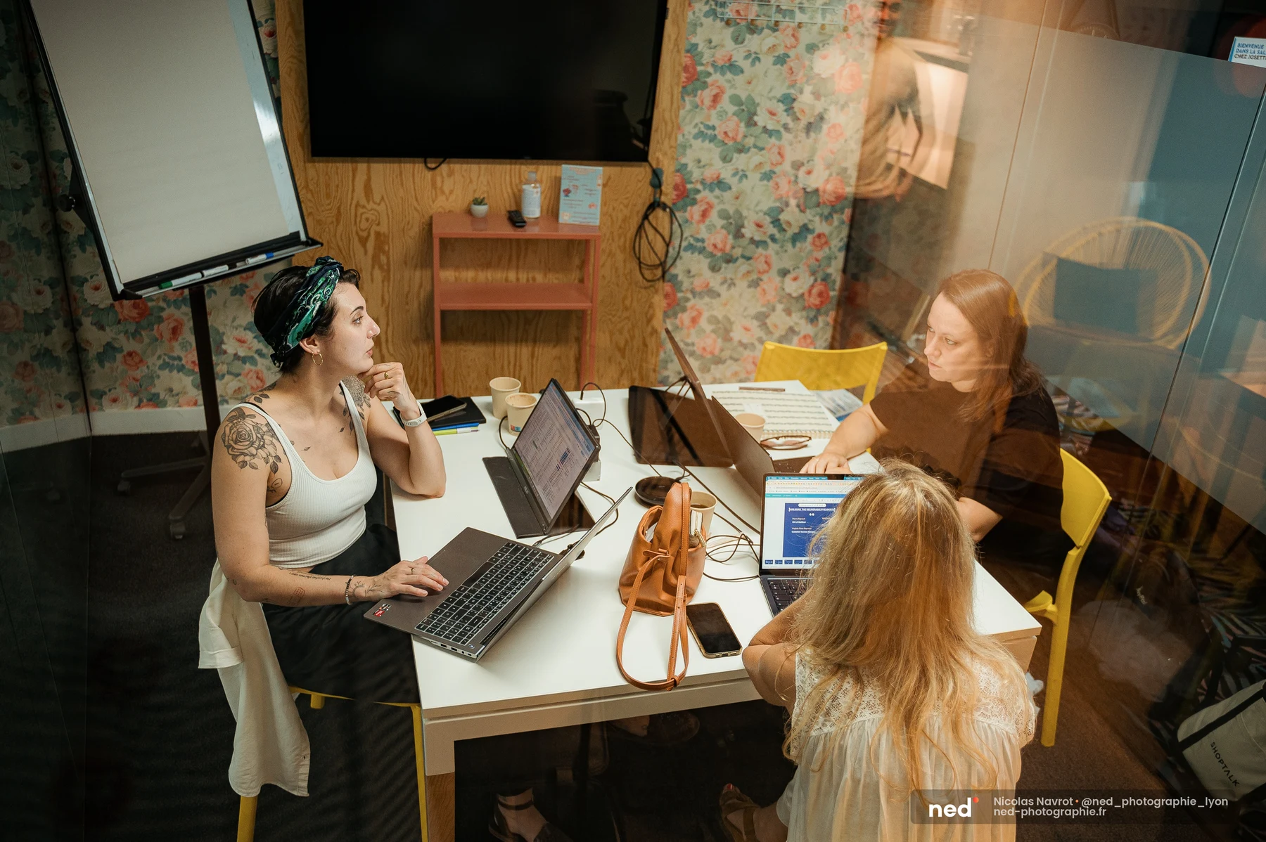 Trois femmes au travail dans une petite salle au papier peint fleuri, laptops ouverts sur une table blanche ; l'une d'elles marque une pause, regard pensif.