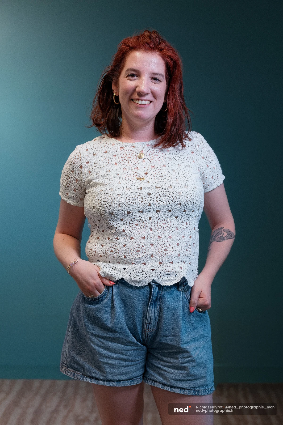 Femme aux cheveux roux mi-longs, debout, mains dans les poches, portant un top blanc en crochet et un short en jean, sur fond vert canard.