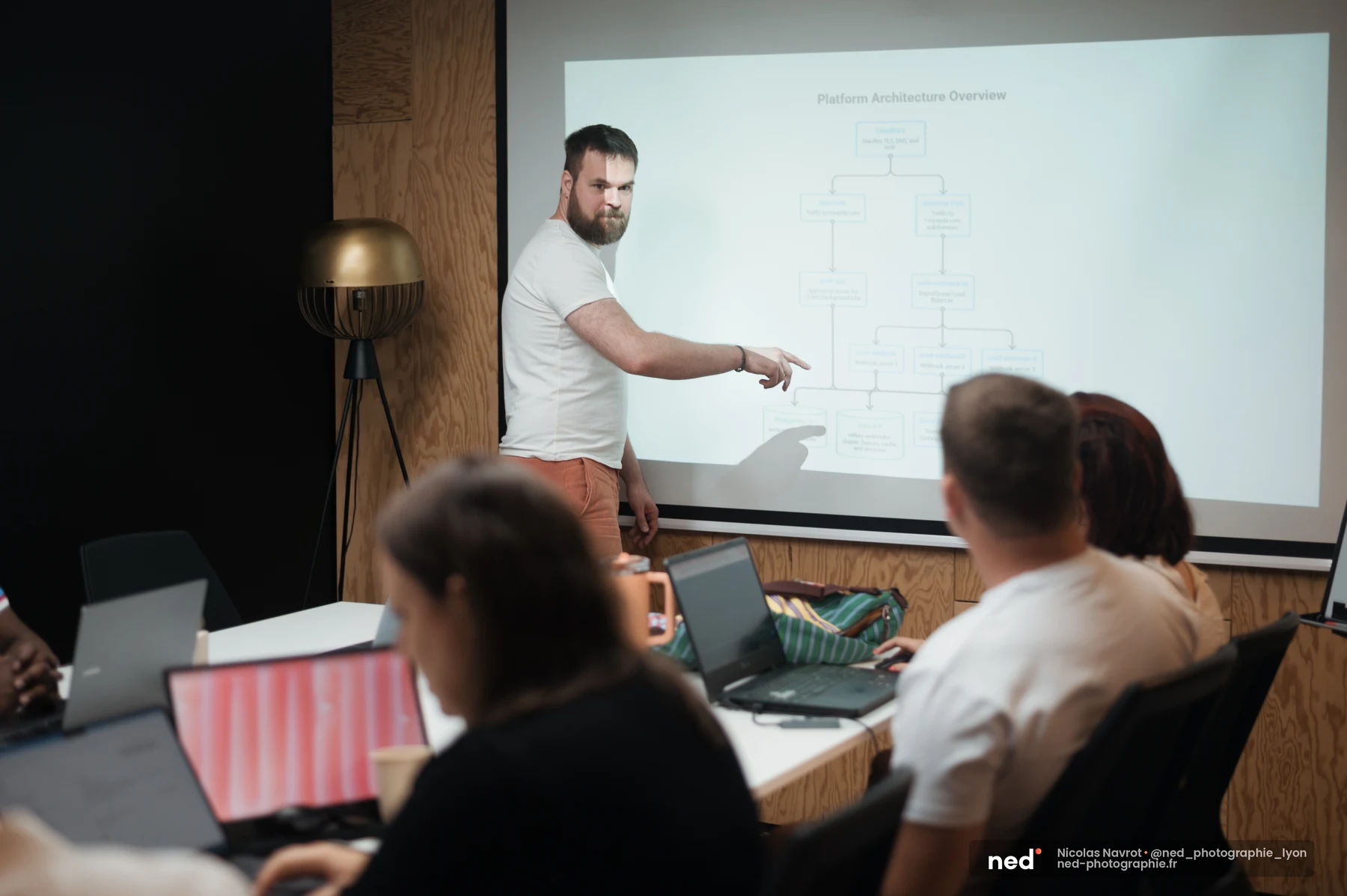Un homme en t-shirt blanc montre du doigt un diagramme d'architecture de plateforme projeté sur un écran, devant un groupe de participants avec laptops.