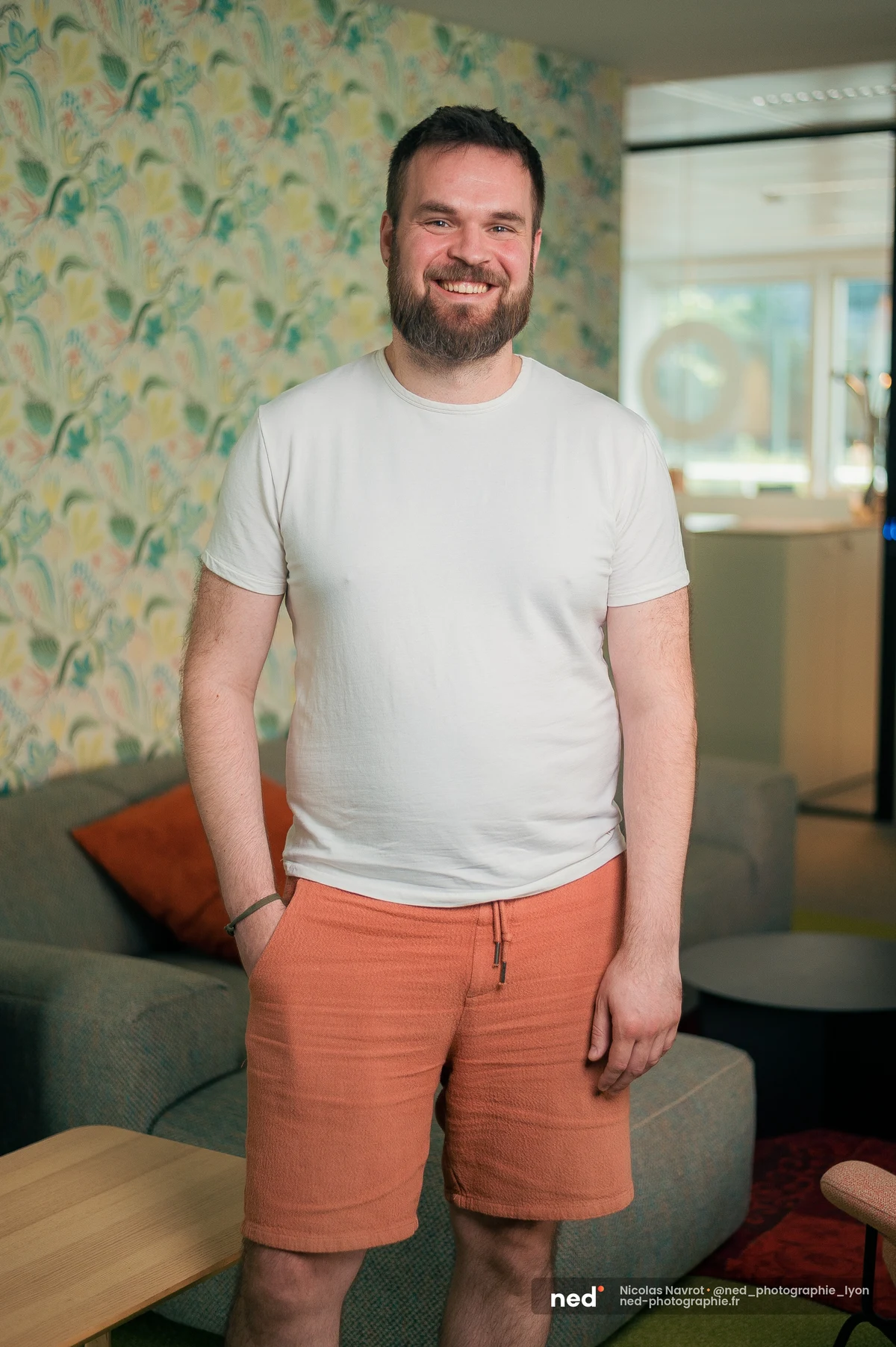 Homme en t-shirt blanc et short terracotta debout dans un espace lounge, devant un mur à motifs floraux colorés.